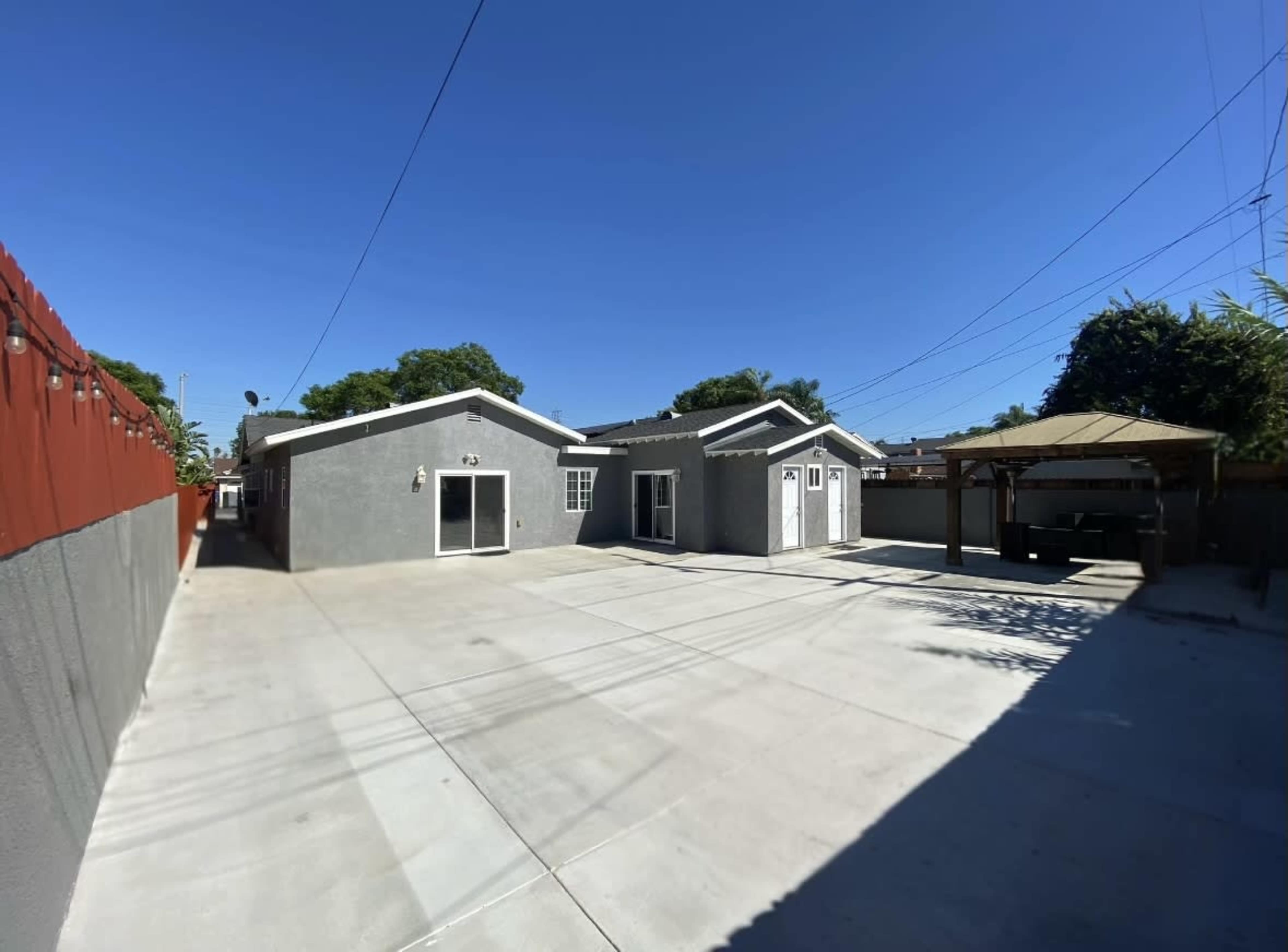 The image shows a spacious concrete yard with two gray buildings and a covered carport in a residential area.