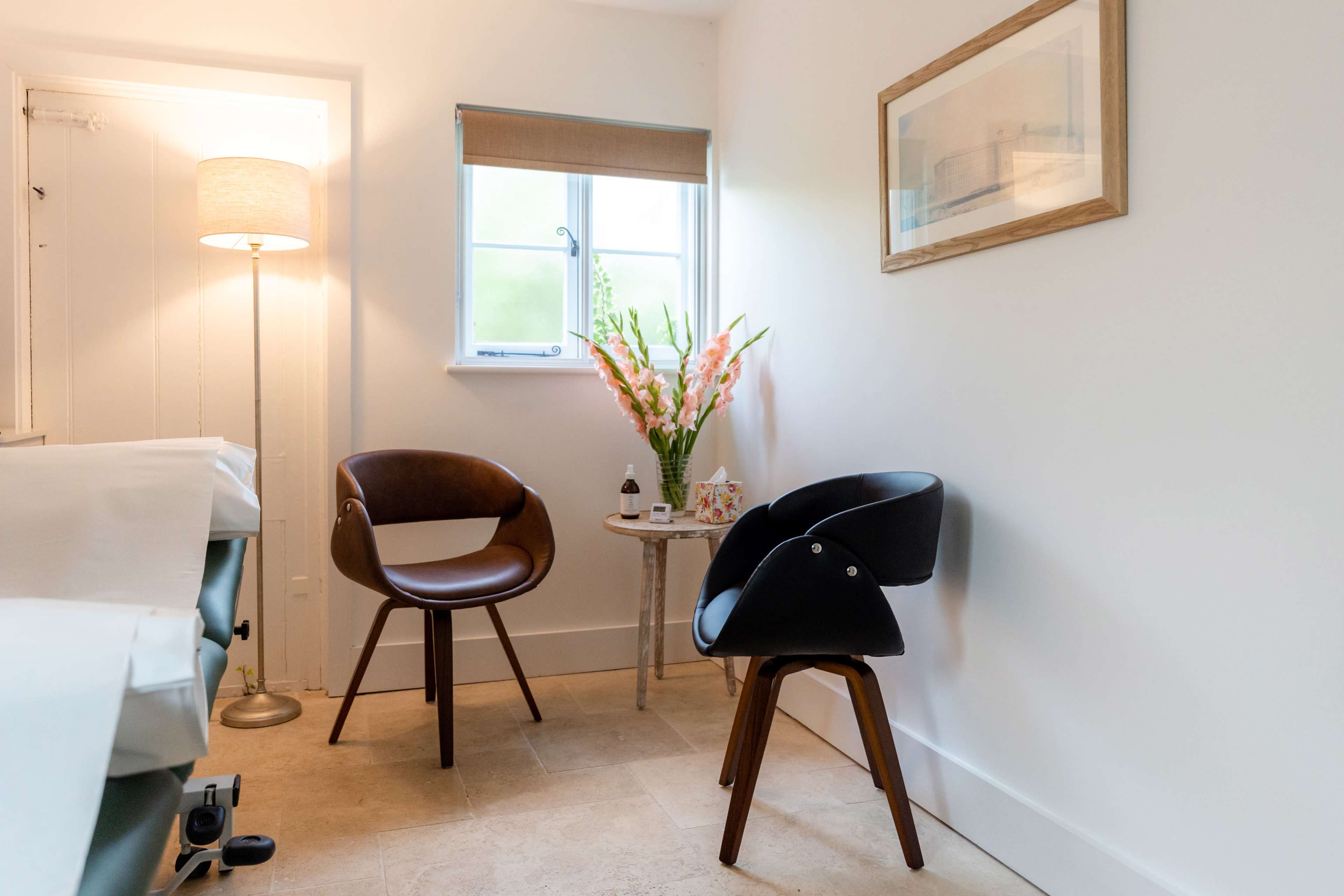 A brightly lit room features two modern chairs beside a small table with floral arrangements and a lamp.