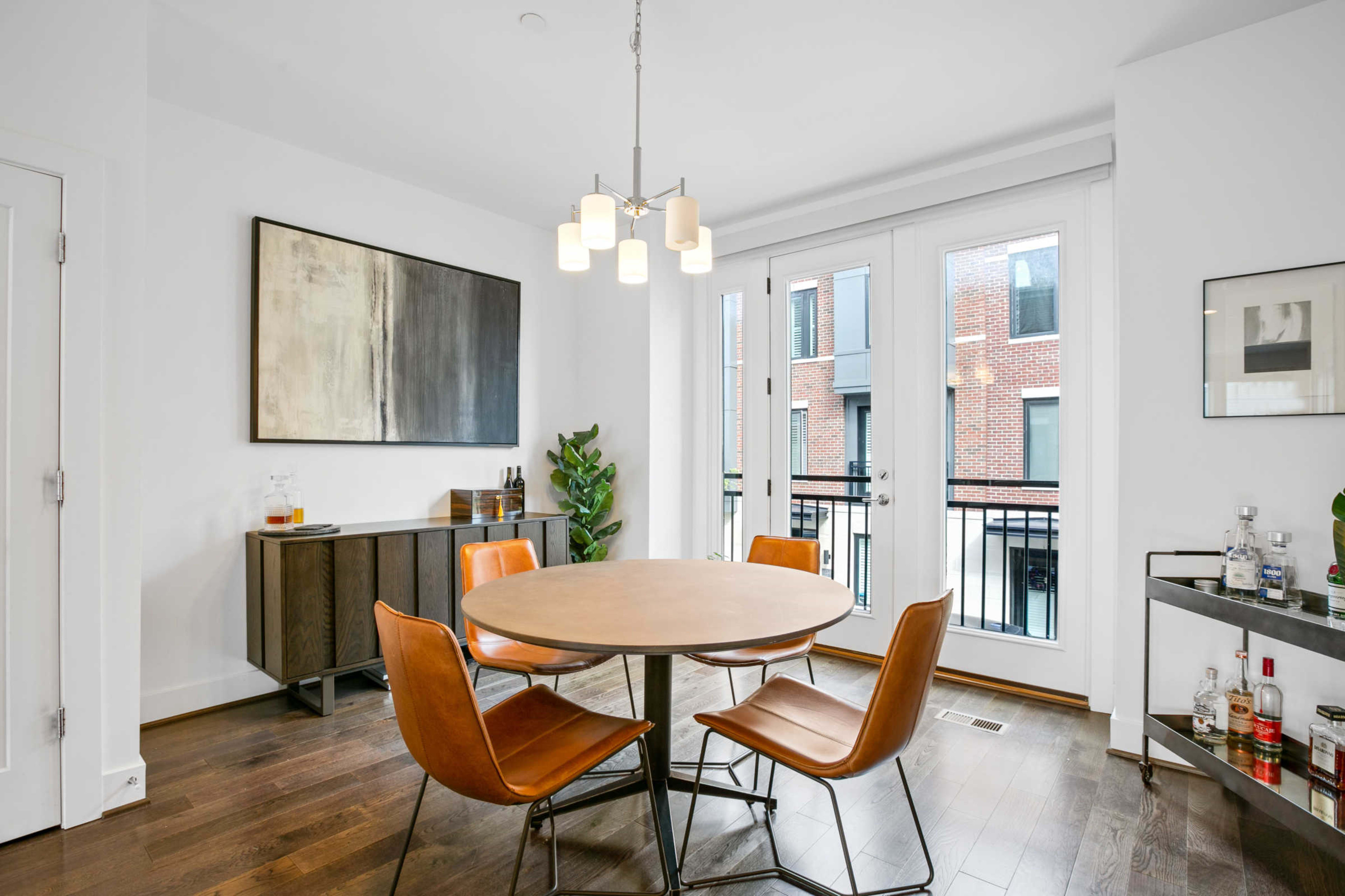 A modern dining area features a round table surrounded by four chairs, with a sideboard against the wall and large windows overlooking a brick exterior.