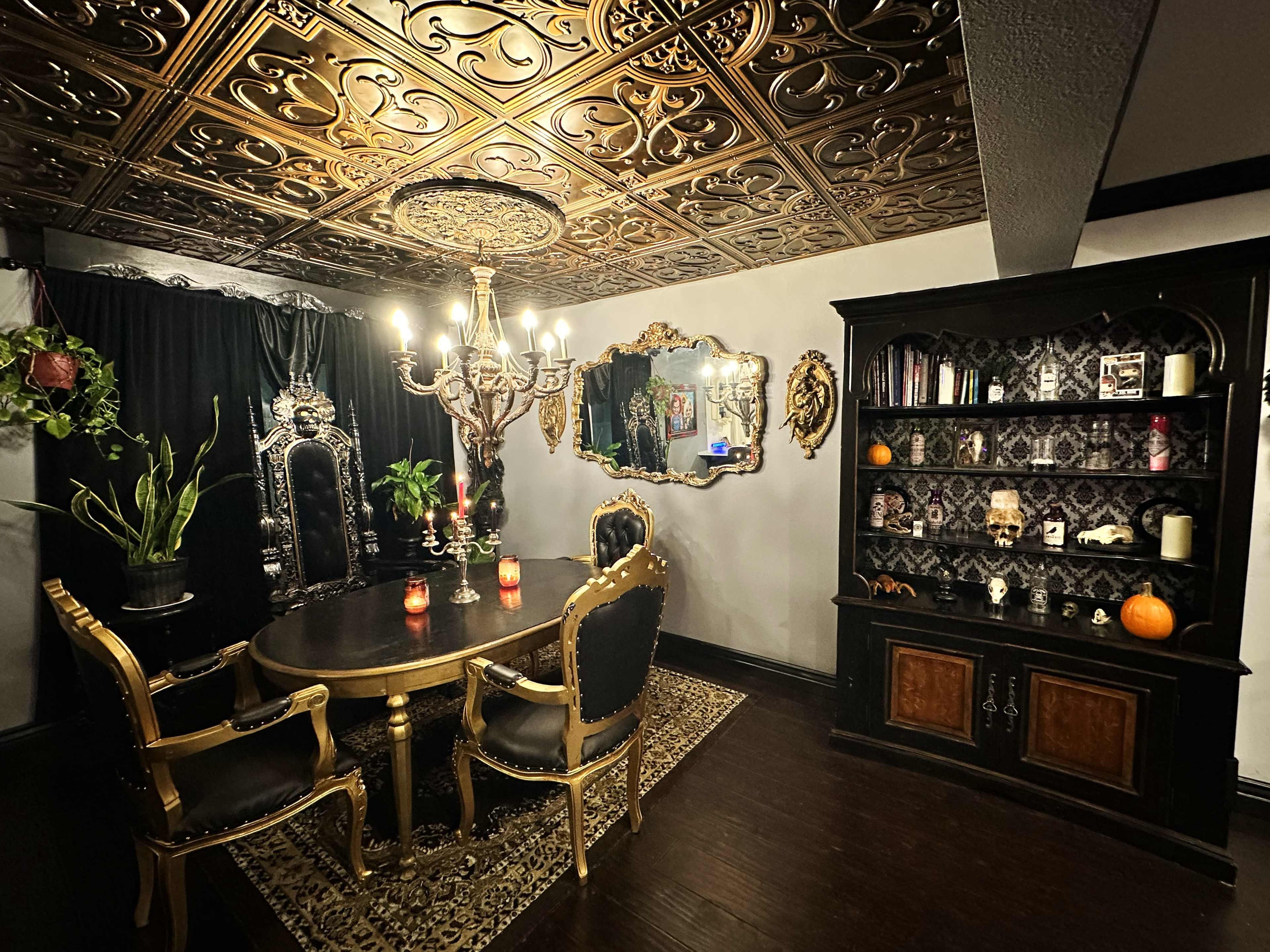 The image shows an elegant dining room featuring a large round table, ornate chairs with gold accents, a decorative chandelier, and a wall adorned with a dark wooden cabinet displaying various decorative items.