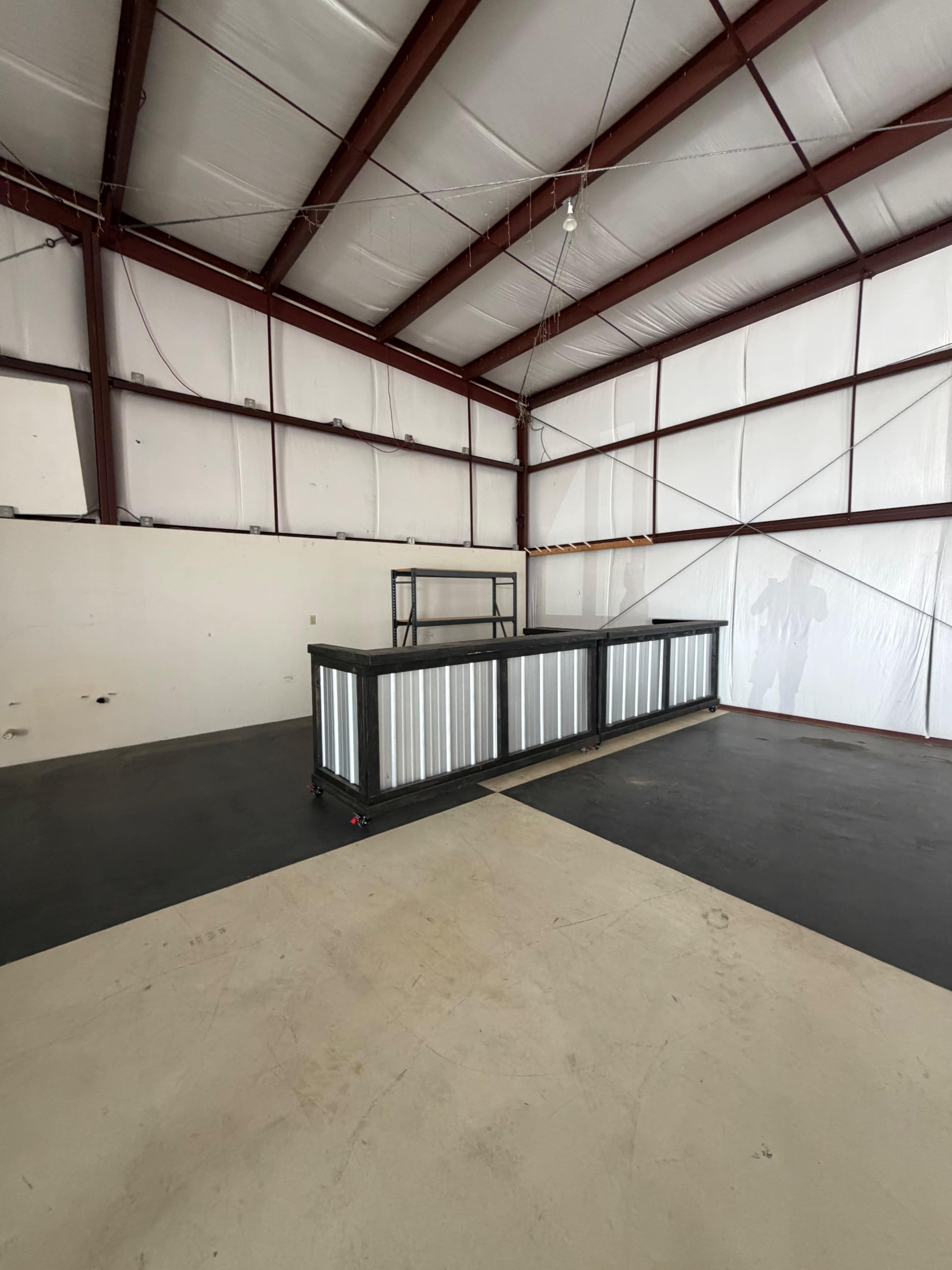 The image shows a large, empty indoor space with a metal counter in the center and walls made of paneling and metal framing.