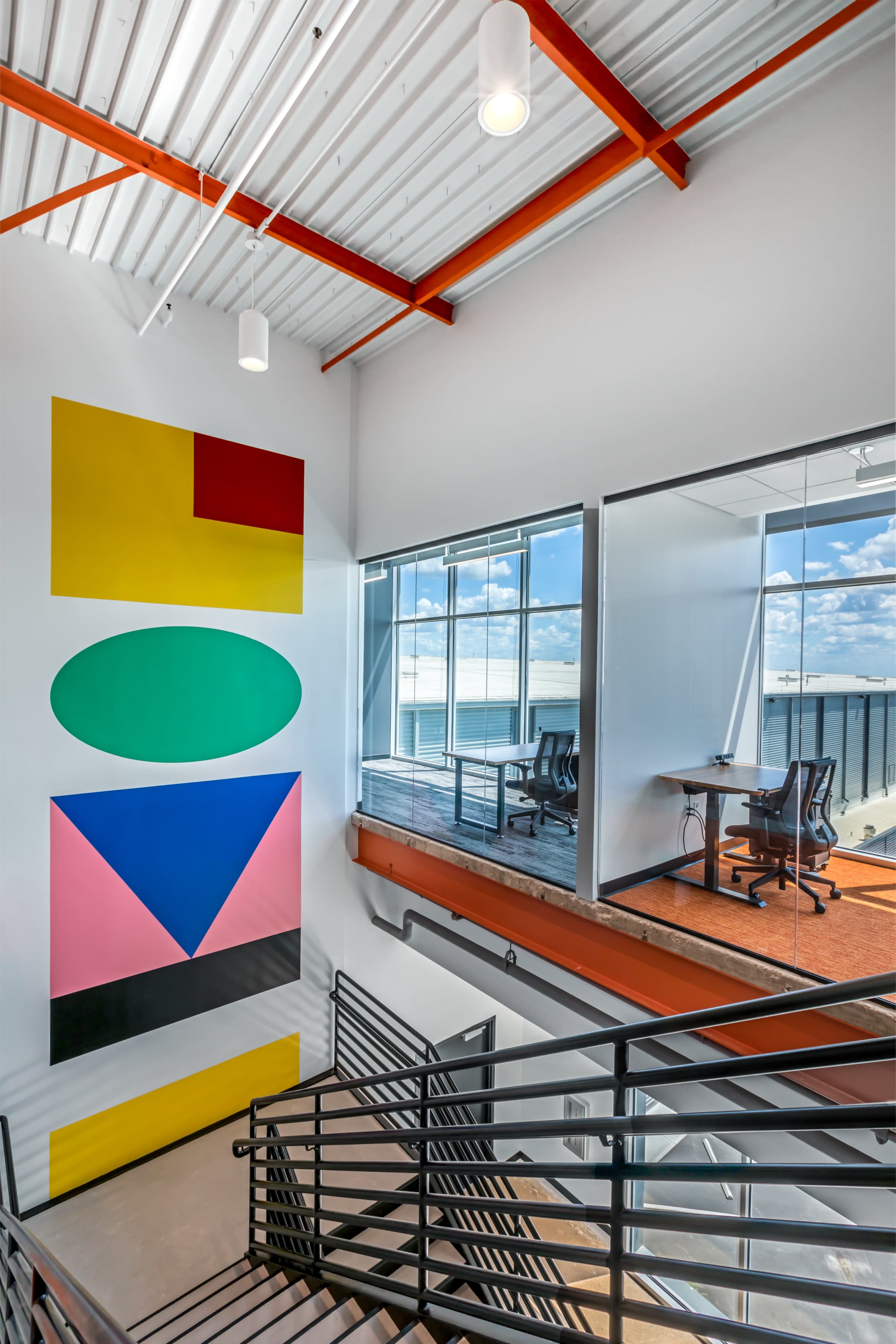 The image shows a modern office interior with a staircase, large windows, and colorful geometric wall art.