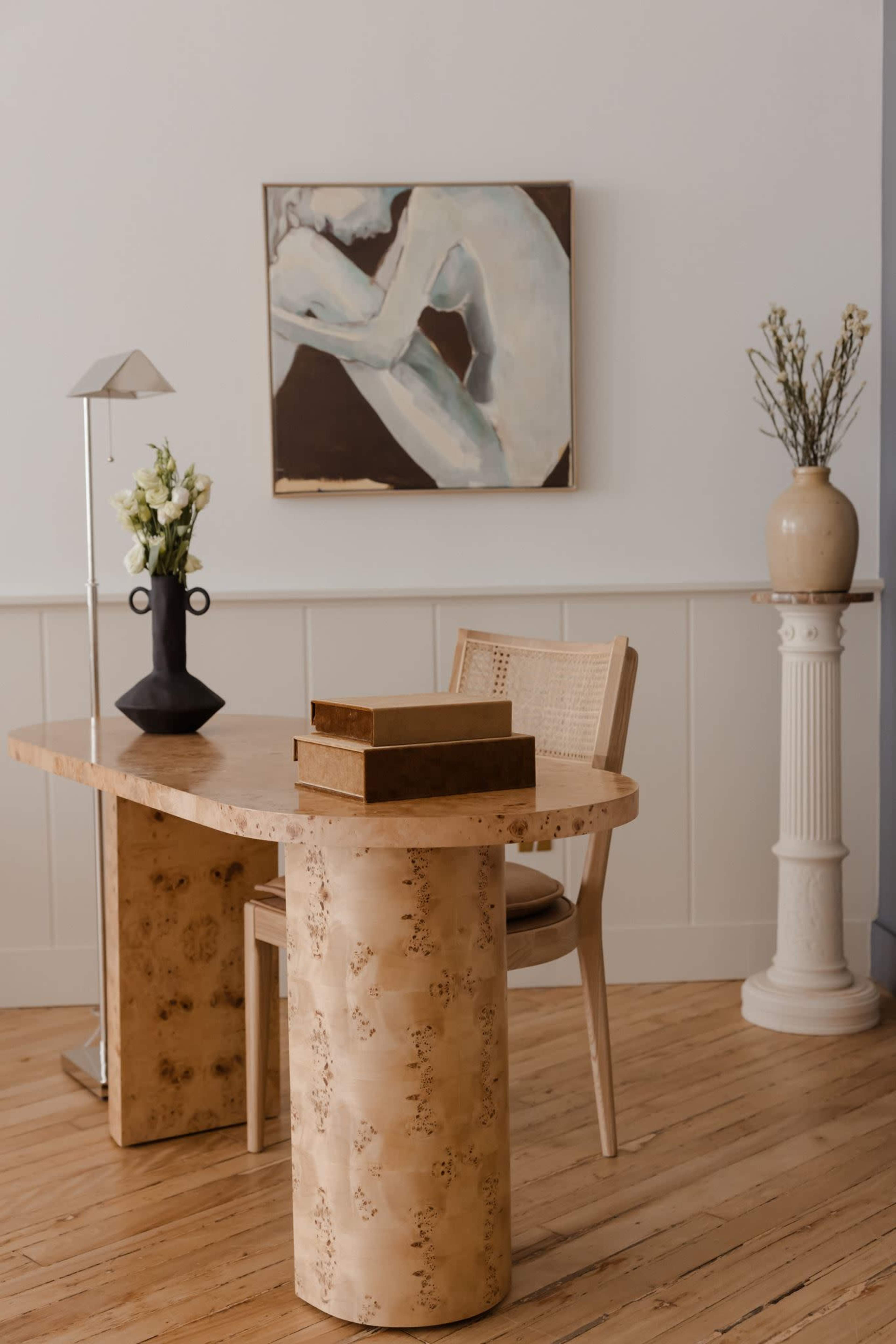 A wooden desk with stacked boxes is positioned in a minimalist room featuring a chair, a lamp, and a large piece of artwork on the wall.