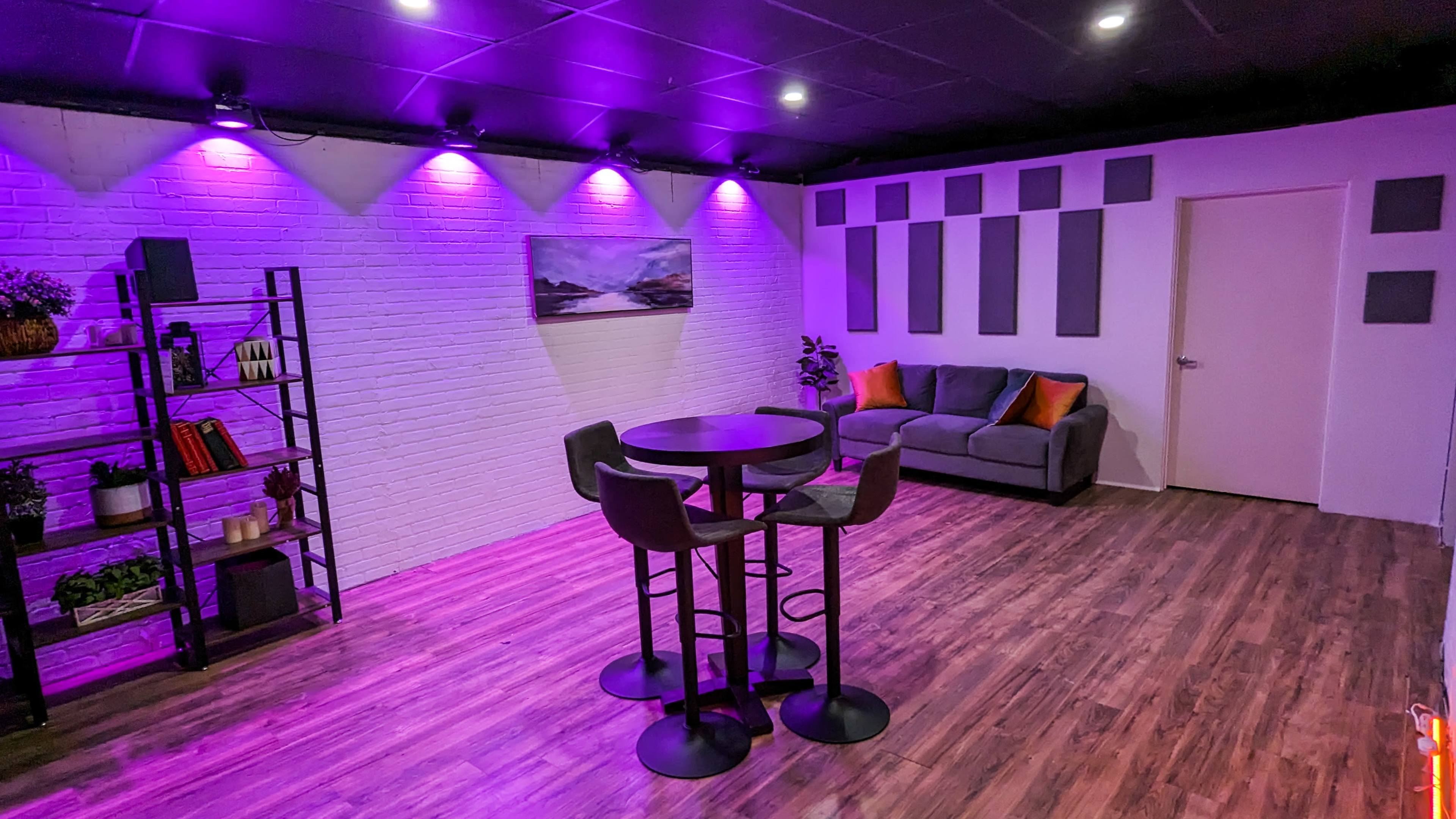 The room features a seating area with a gray couch, a round table surrounded by bar stools, and purple accent lighting illuminating the walls.