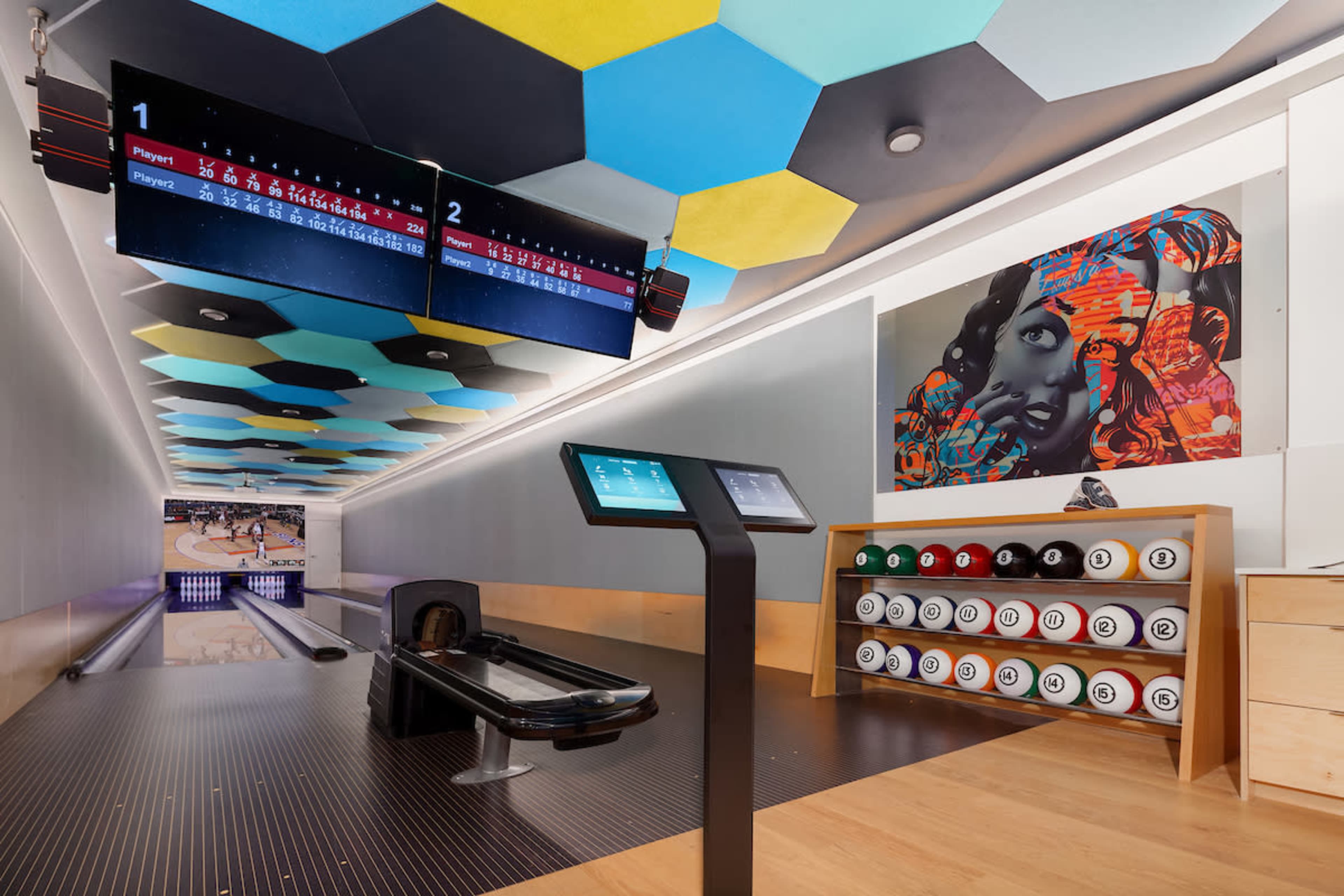 A modern bowling alley featuring a digital score display, a bowling lane with an automated ball return system, and colorful hexagonal ceiling tiles, along with a mural and a rack of bowling balls on the wall.