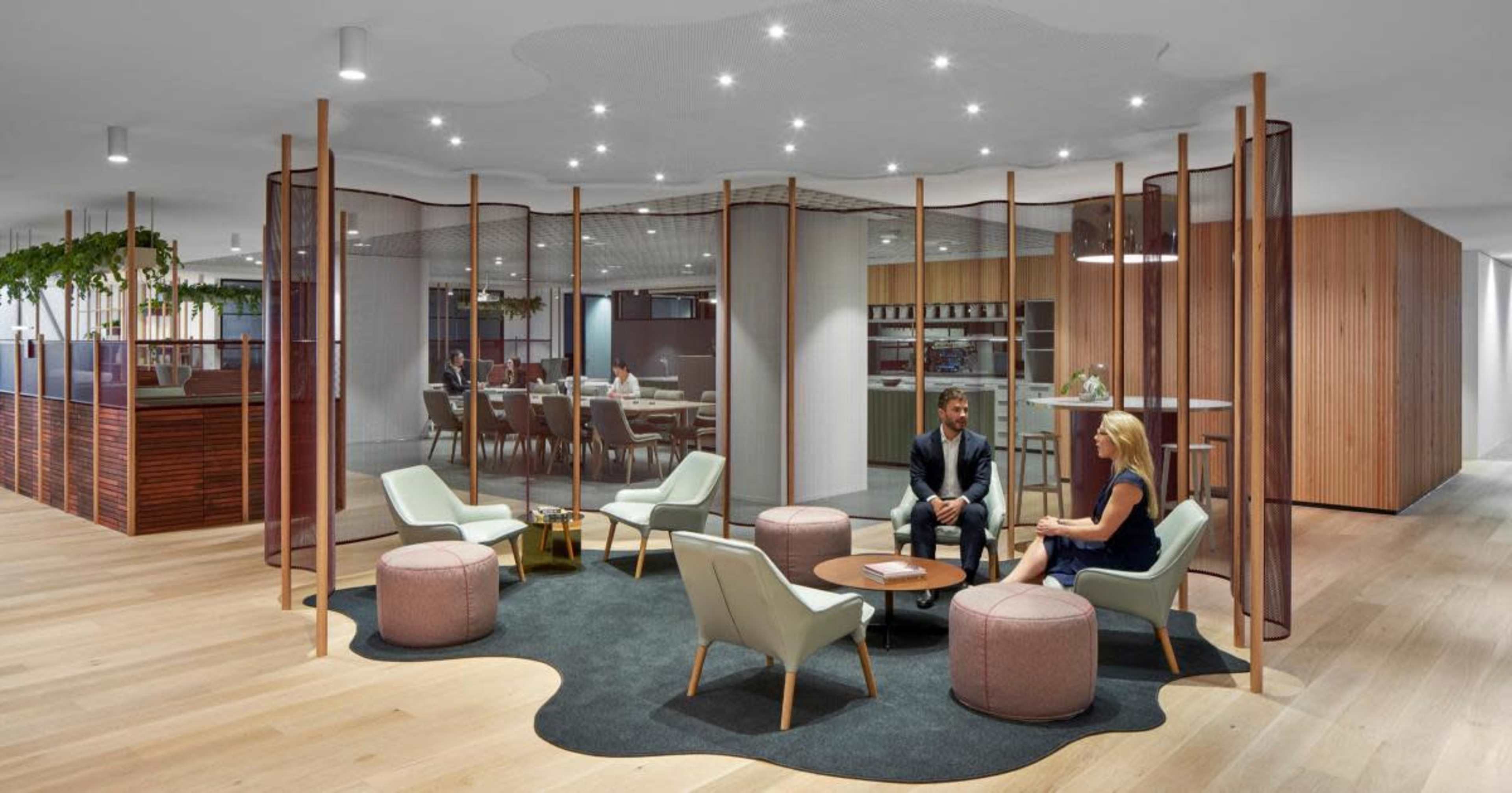 The image shows a modern office space with a seating area featuring light-colored furniture, a circular table, and wooden partitions separating different work areas.