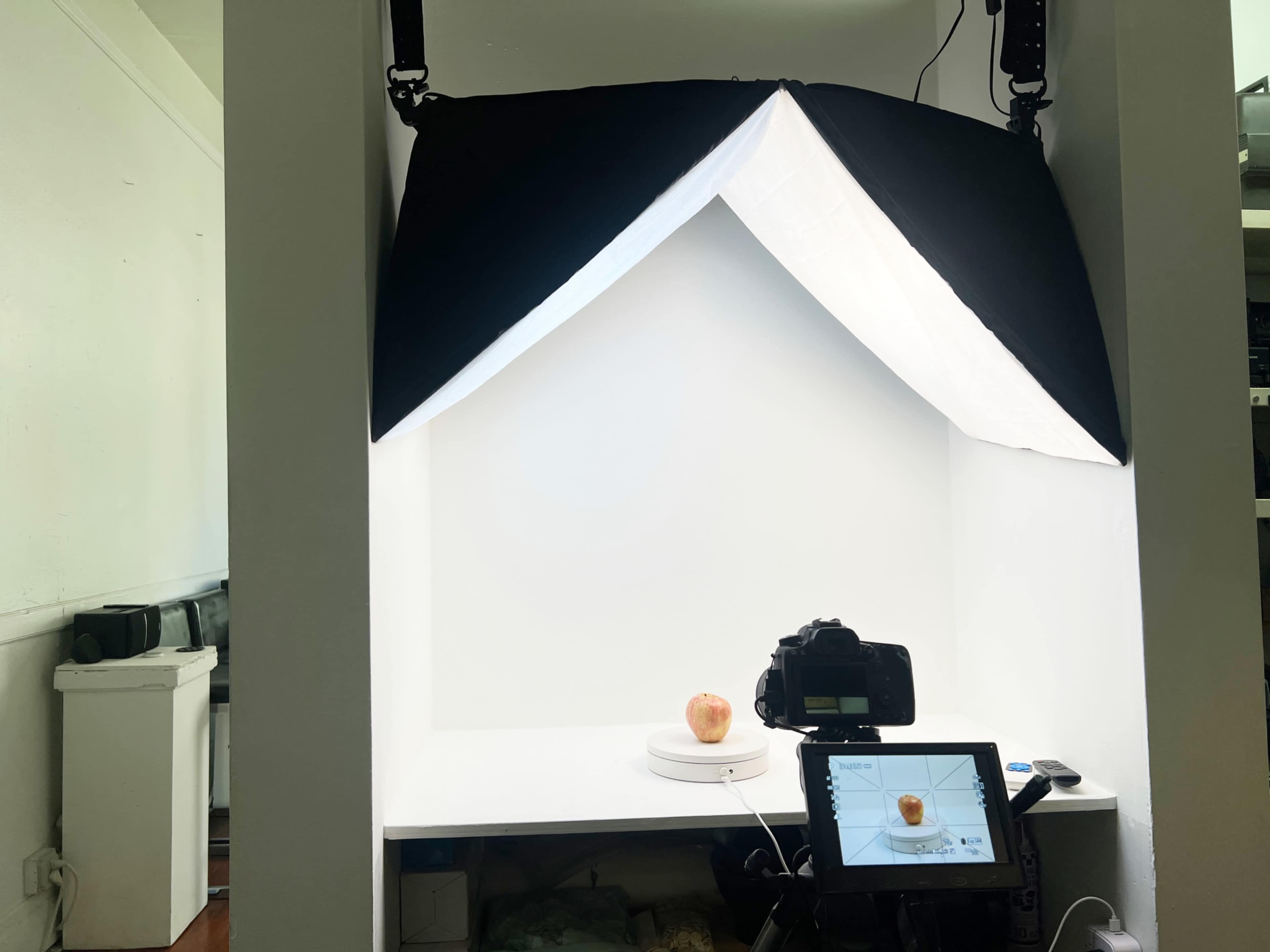 A photography setup features an isolated apple on a turntable under softbox lighting, with a camera and monitor positioned nearby.
