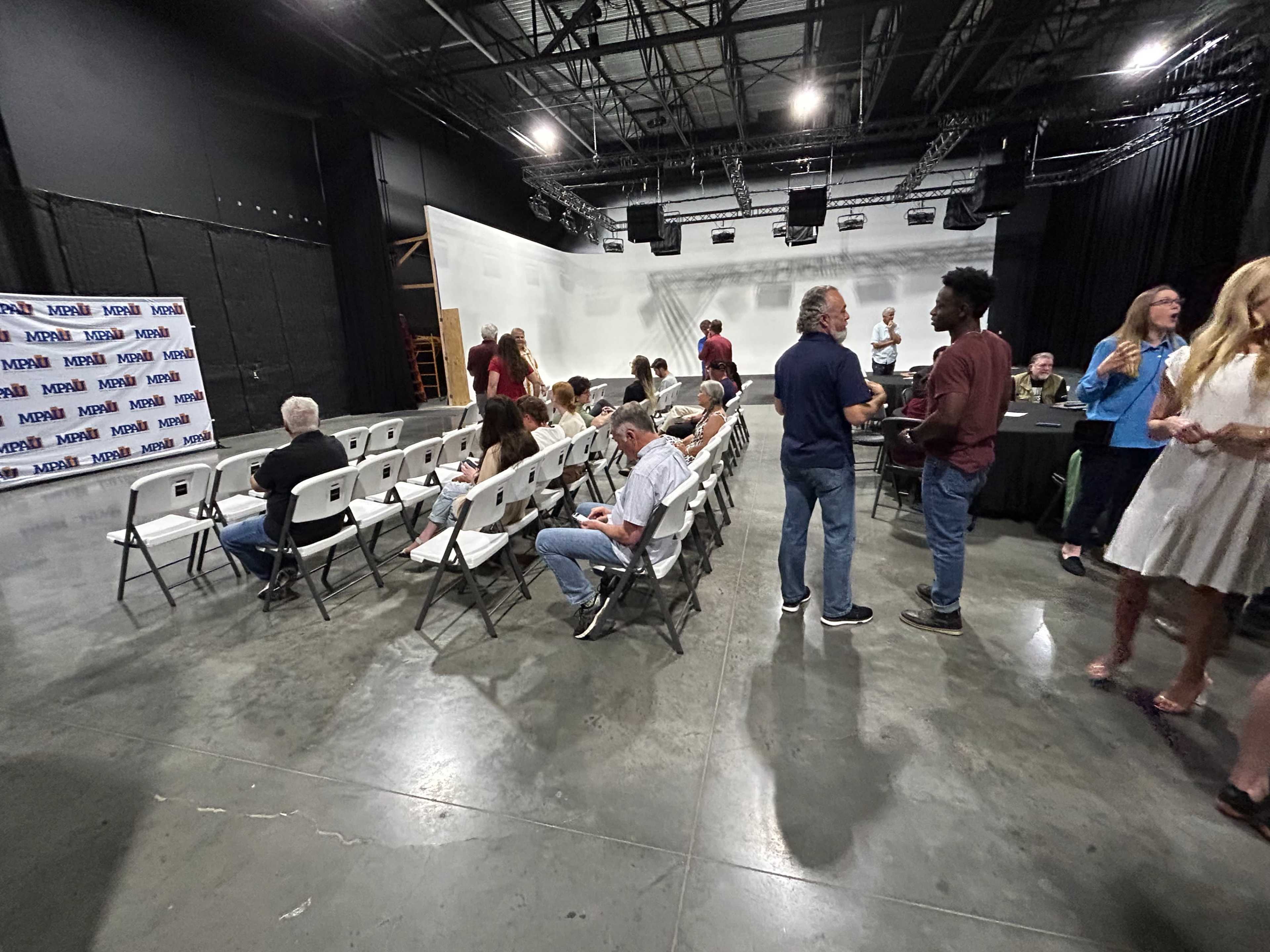 A semi-filled auditorium with rows of white folding chairs and people gathered, some seated and others standing near a black table.