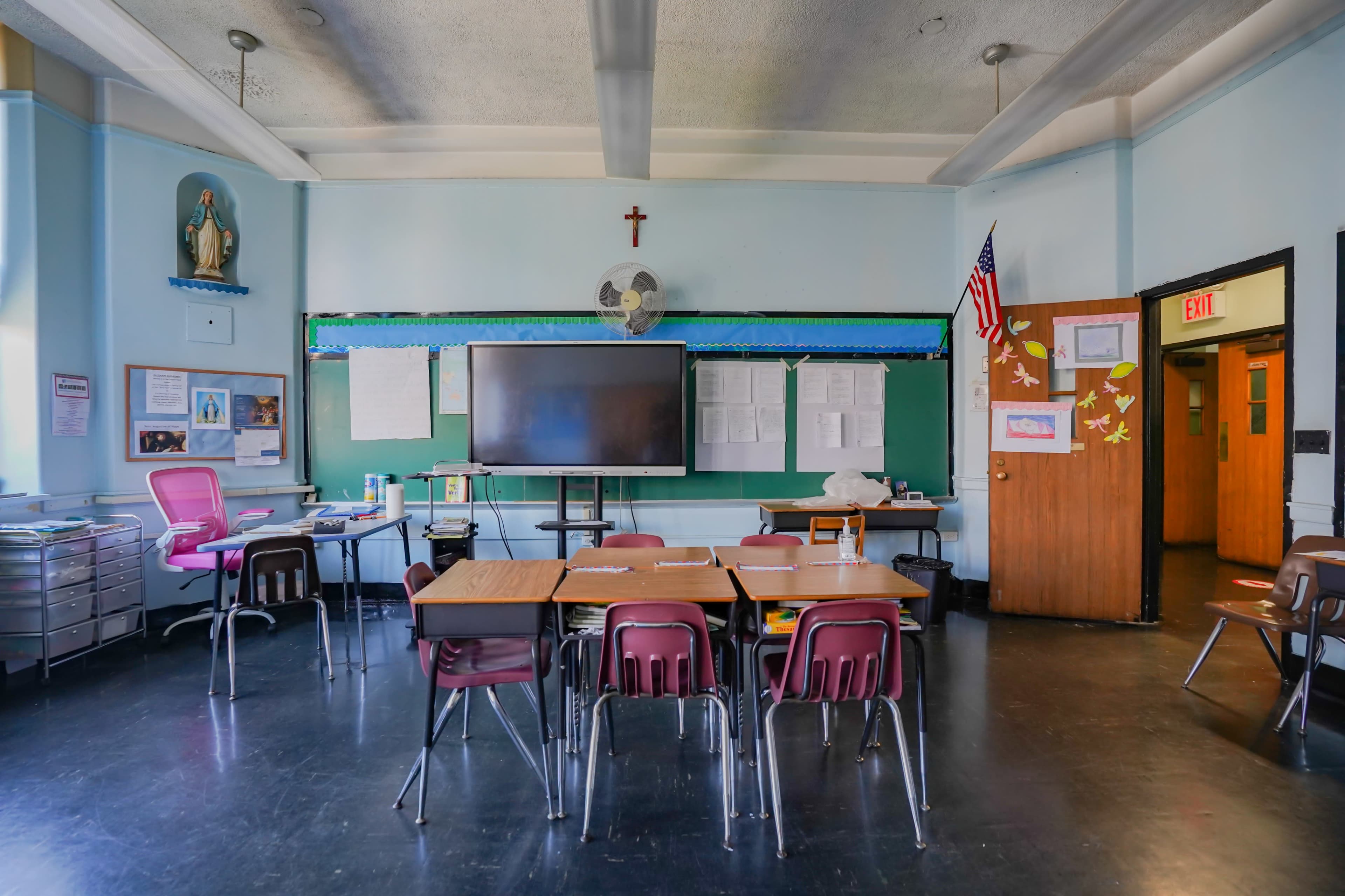 A classroom with desks arranged in a central area, a large screen on the front wall, posters, and a door leading outside.