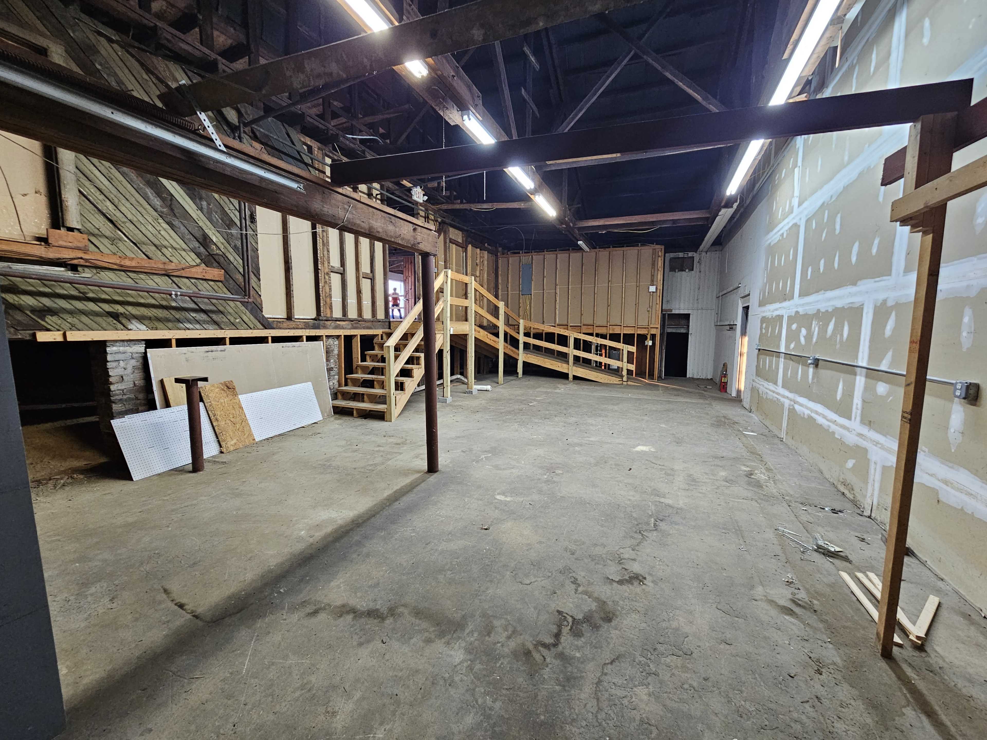 The image shows an empty, spacious warehouse interior with wooden beams, a staircase, and unfinished walls.