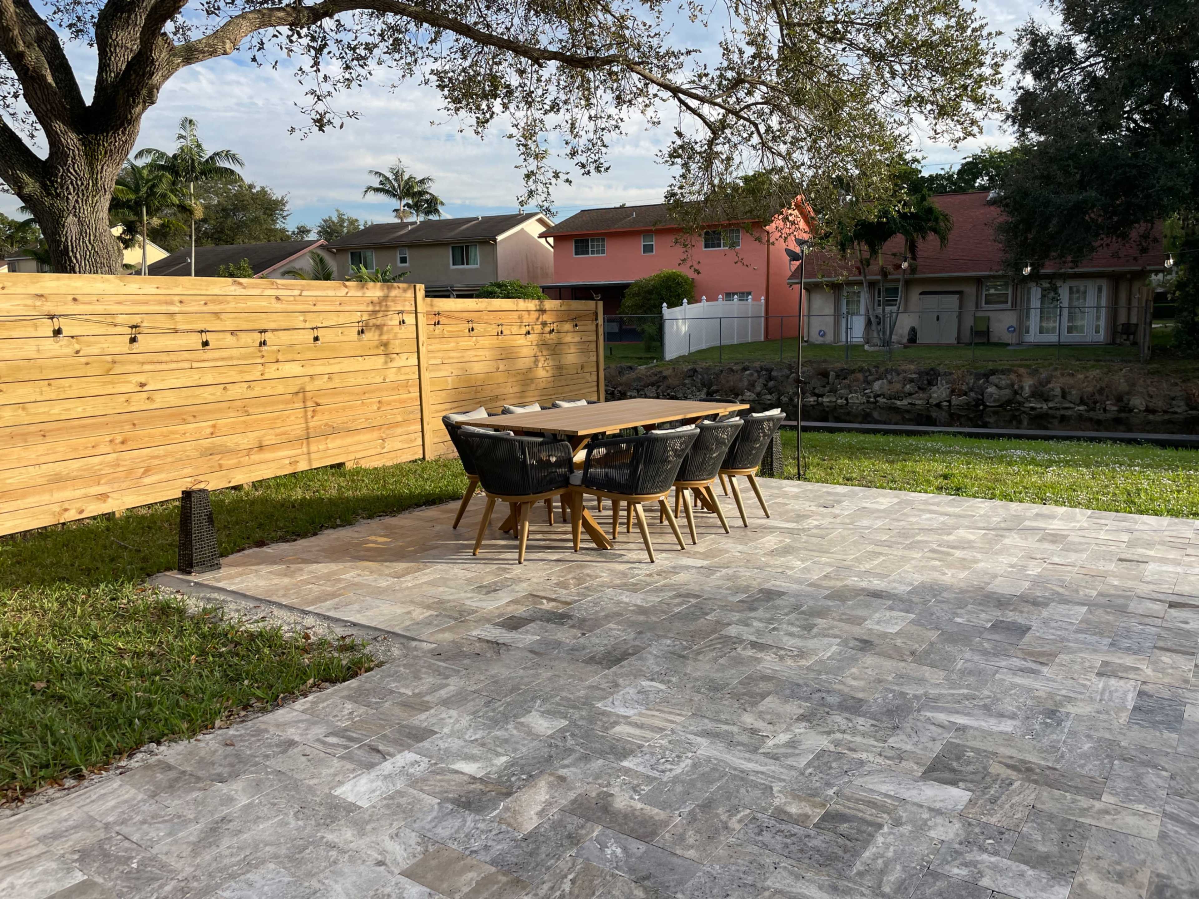 A wooden dining table with black chairs is set up on a stone patio beside a grassy area and a canal, with a wooden fence in the background.