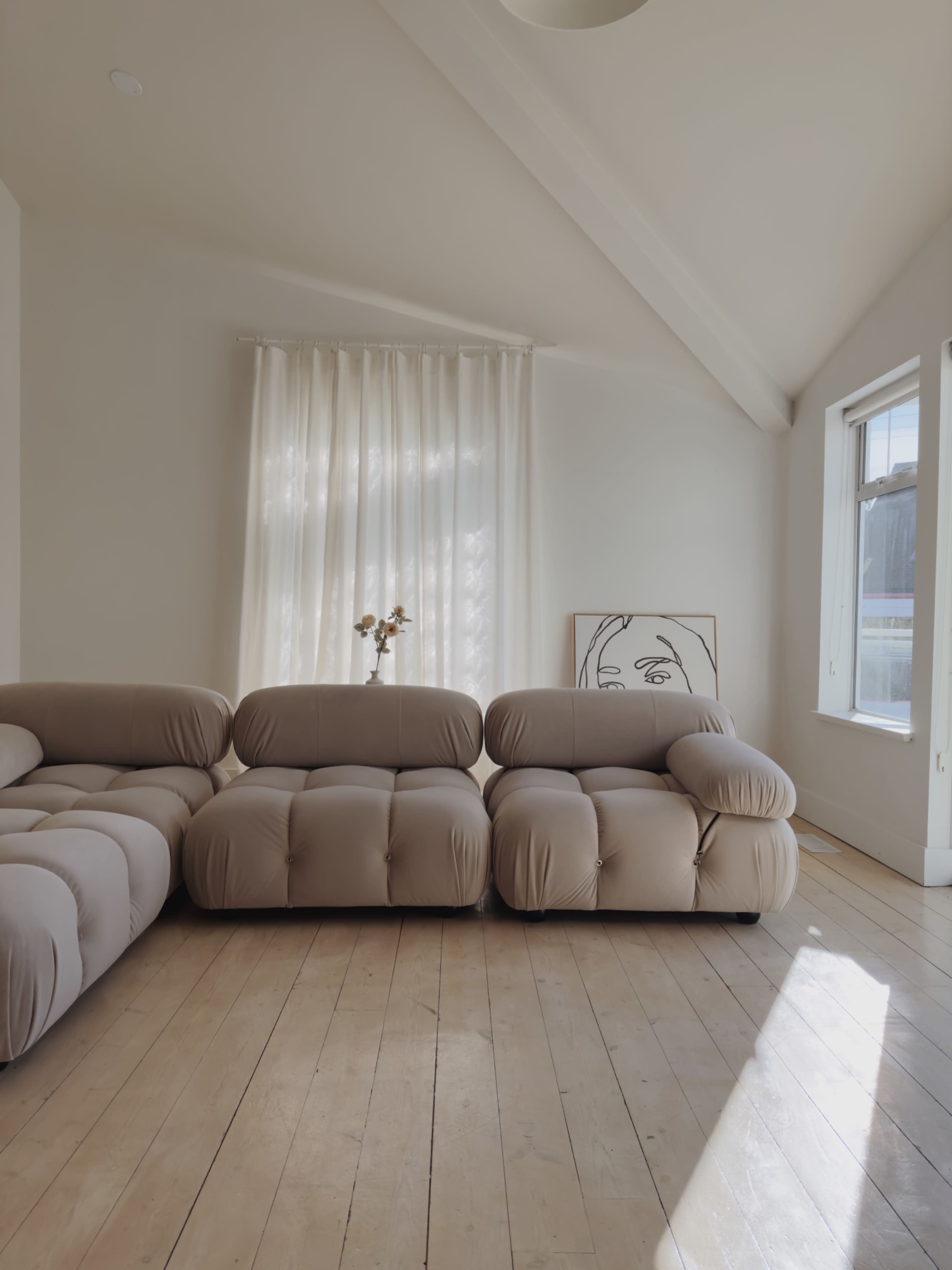 A light-filled living space features a beige, bulbous sofa, wooden floorboards, and sheer curtains beside a framed artwork.