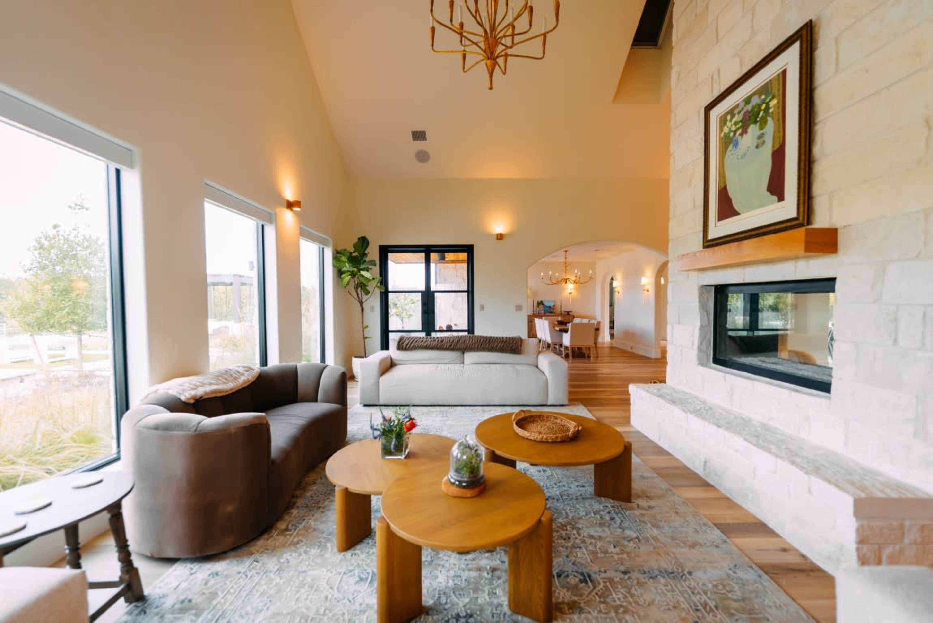 The image shows a cozy living room with large windows, a fireplace, and modern furniture arranged around wooden coffee tables.