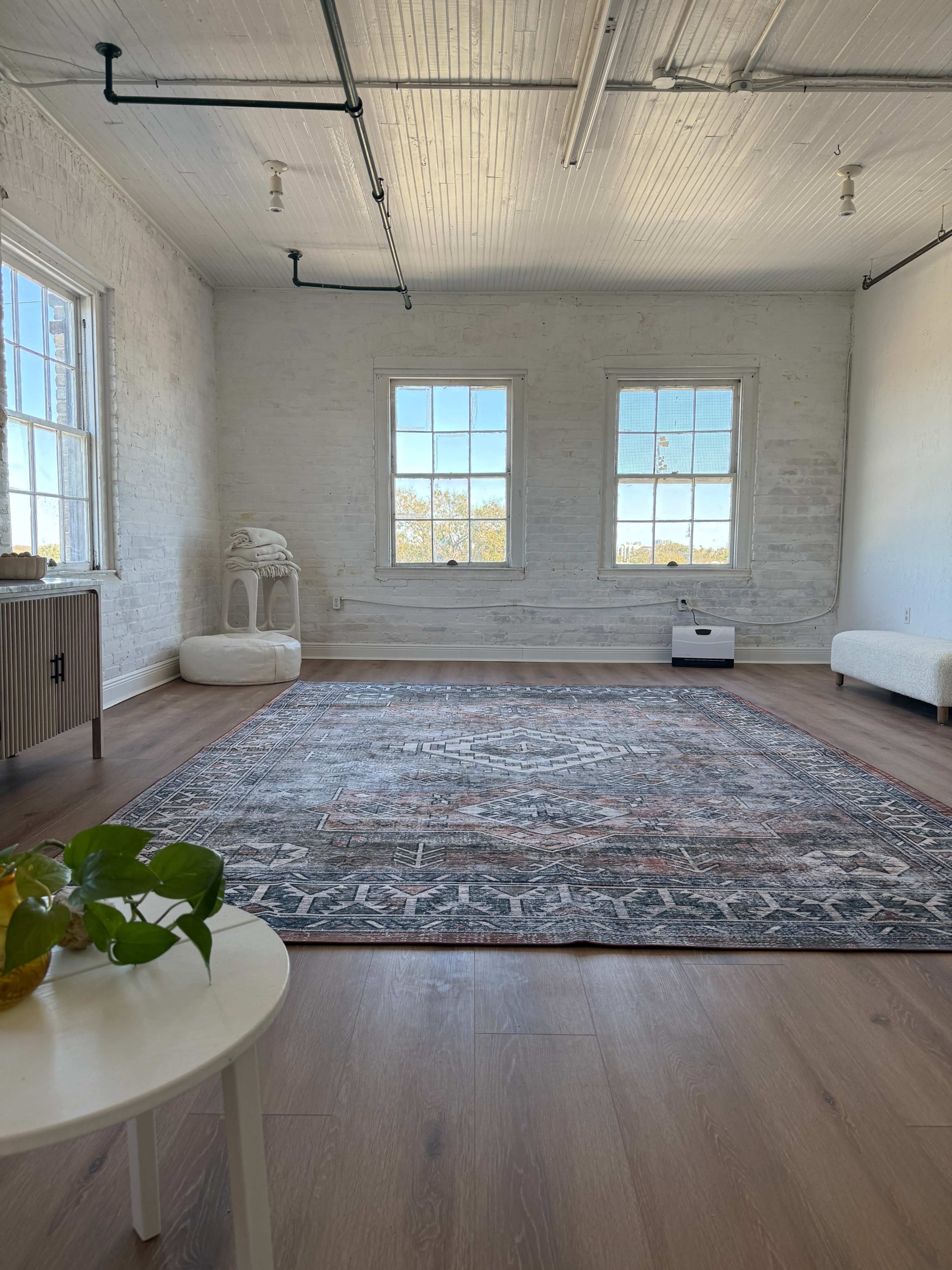 The image shows a spacious room with white brick walls, two windows letting in natural light, a patterned rug on the wooden floor, and minimal furnishings.