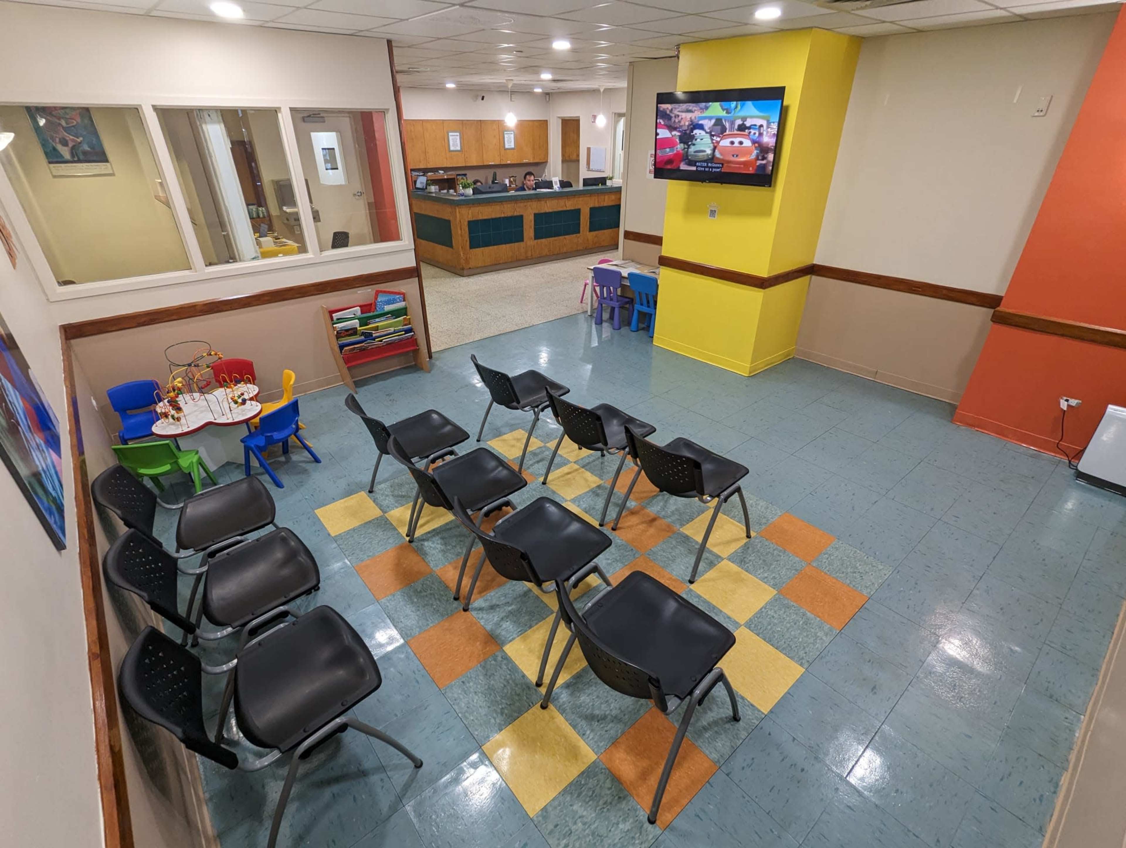 The image shows a waiting room with black chairs arranged in rows, colorful tile flooring, and a reception area visible in the background with a television displaying a cartoon.