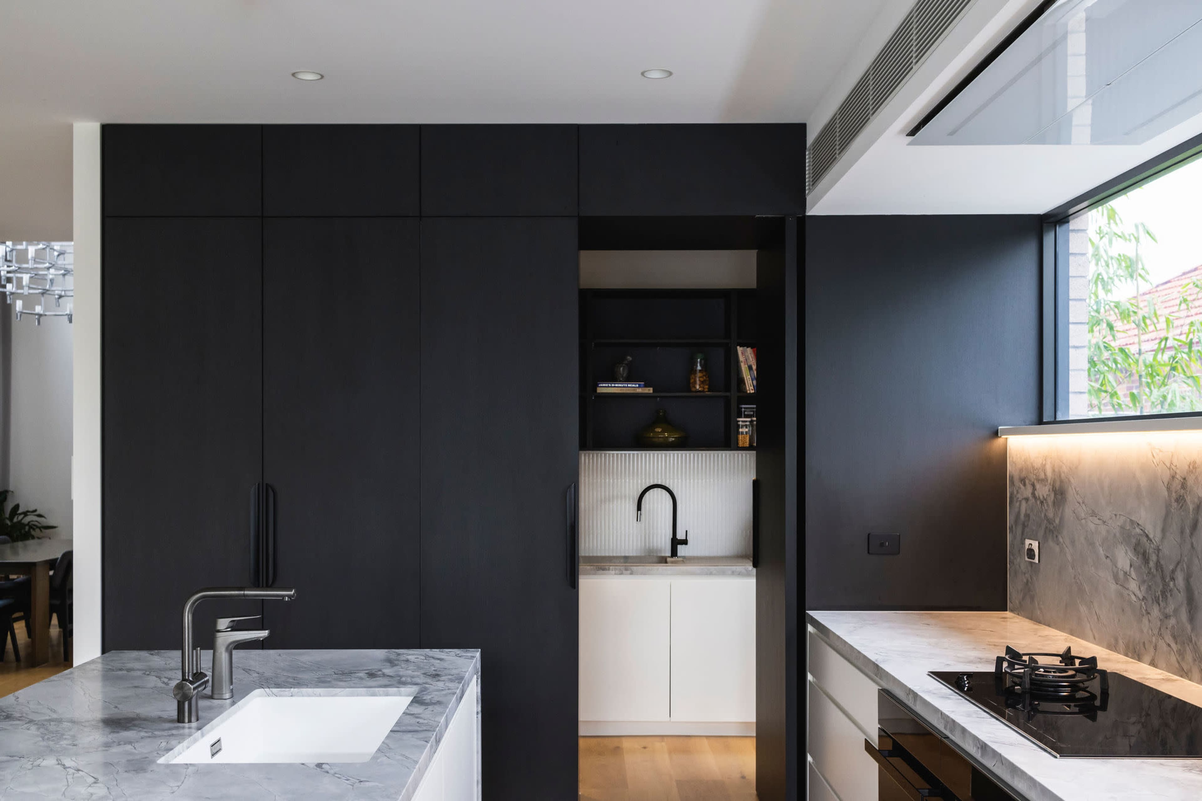 A modern kitchen with sleek black cabinetry, a marble countertop, and a small, open pantry area visible in the background.