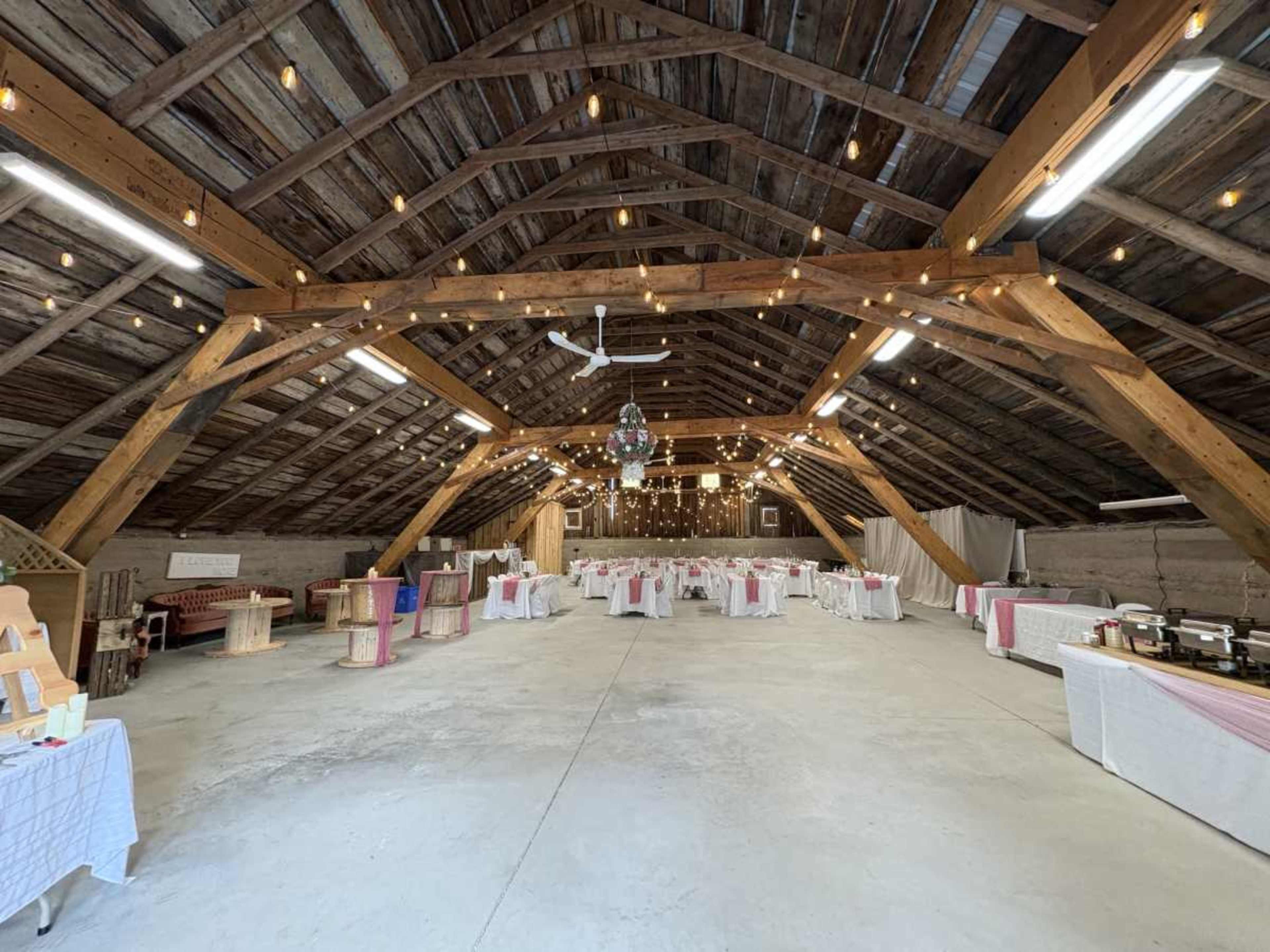 The image shows a spacious, rustic hall with wooden beams, dining tables covered in white and pink linens, and string lights hanging from the ceiling.