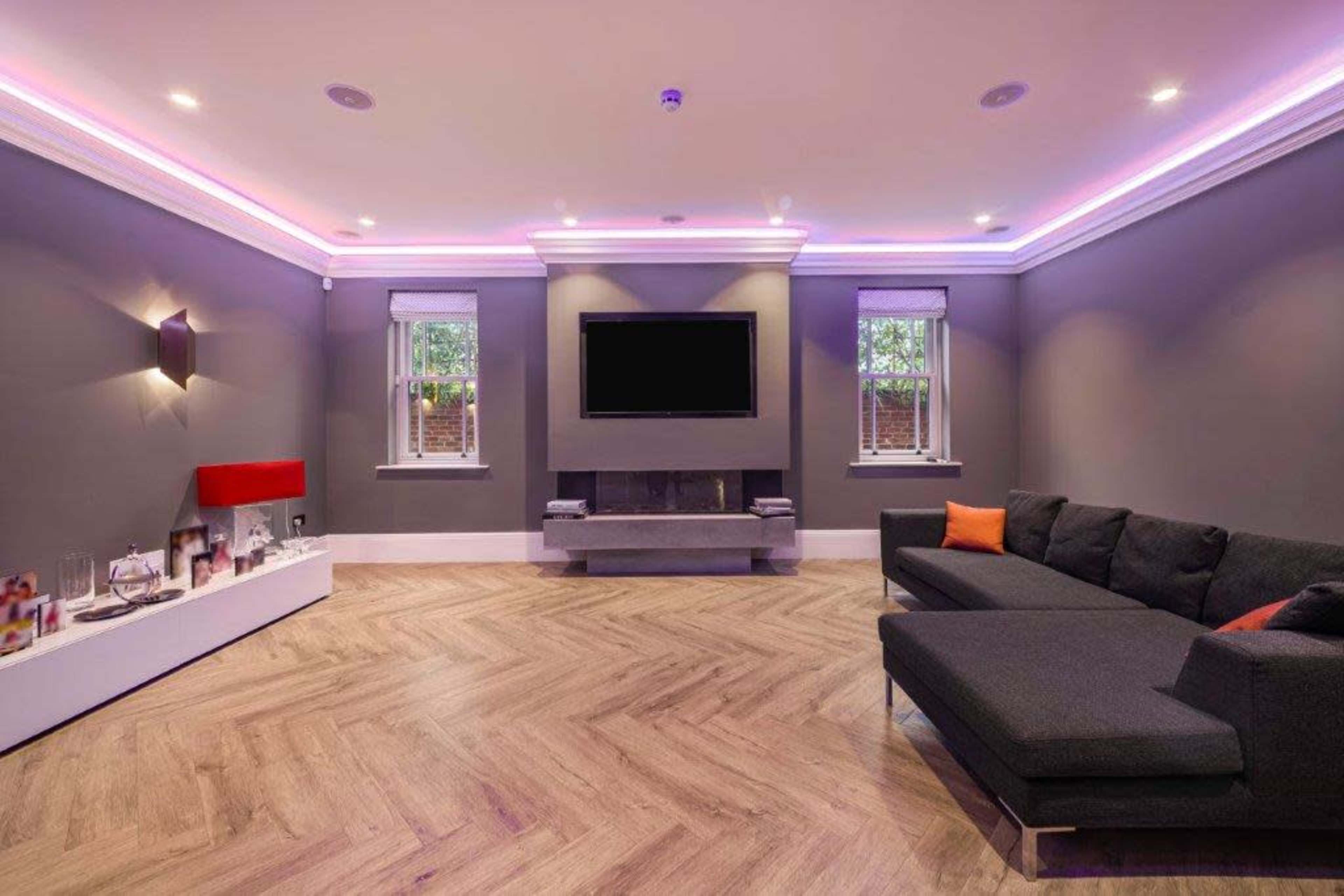 The image shows a modern living room with gray walls, a large sectional sofa, a flat-screen TV mounted on the wall, and ambient lighting.
