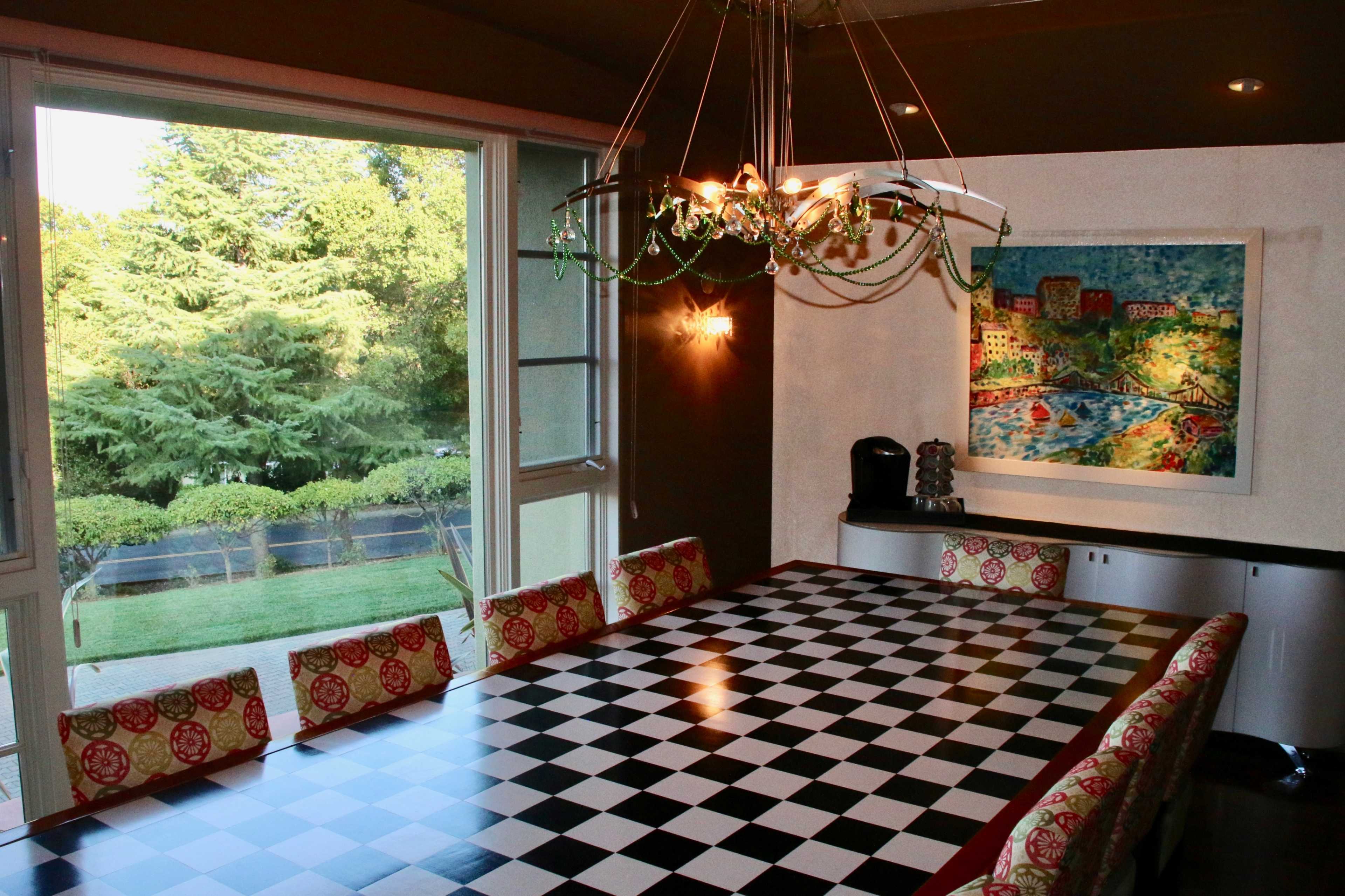 A large dining table with a checkered pattern is set in a well-lit room featuring a window that offers a view of greenery outside.