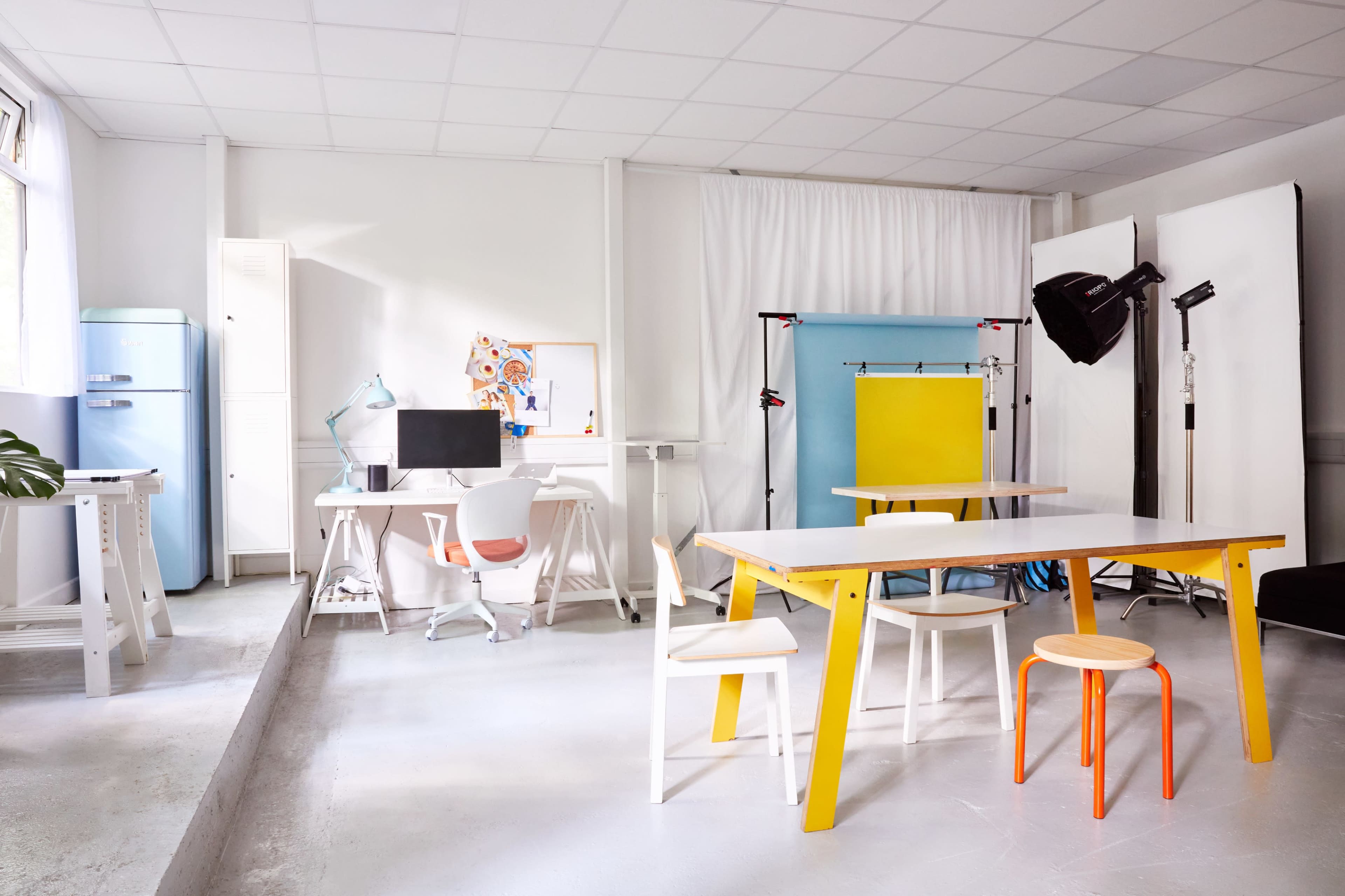 A bright and minimalistic studio space with various furniture pieces, including a yellow table, white chairs, and a blue background setup for photography.