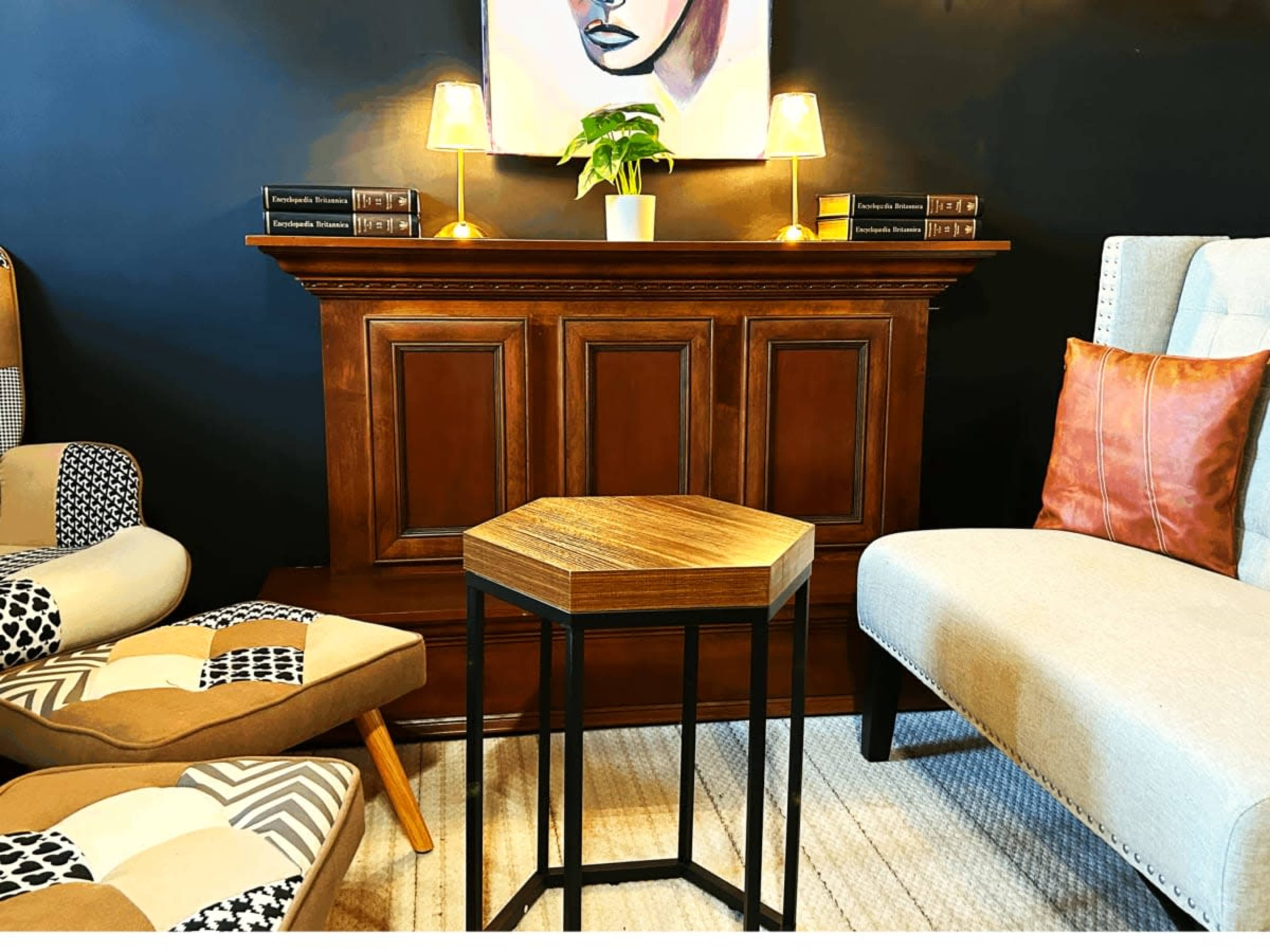 The image shows a cozy seating area featuring a patterned armchair, a light-colored sofa, a small hexagonal table, and a wooden credenza with lamps and a potted plant on top.