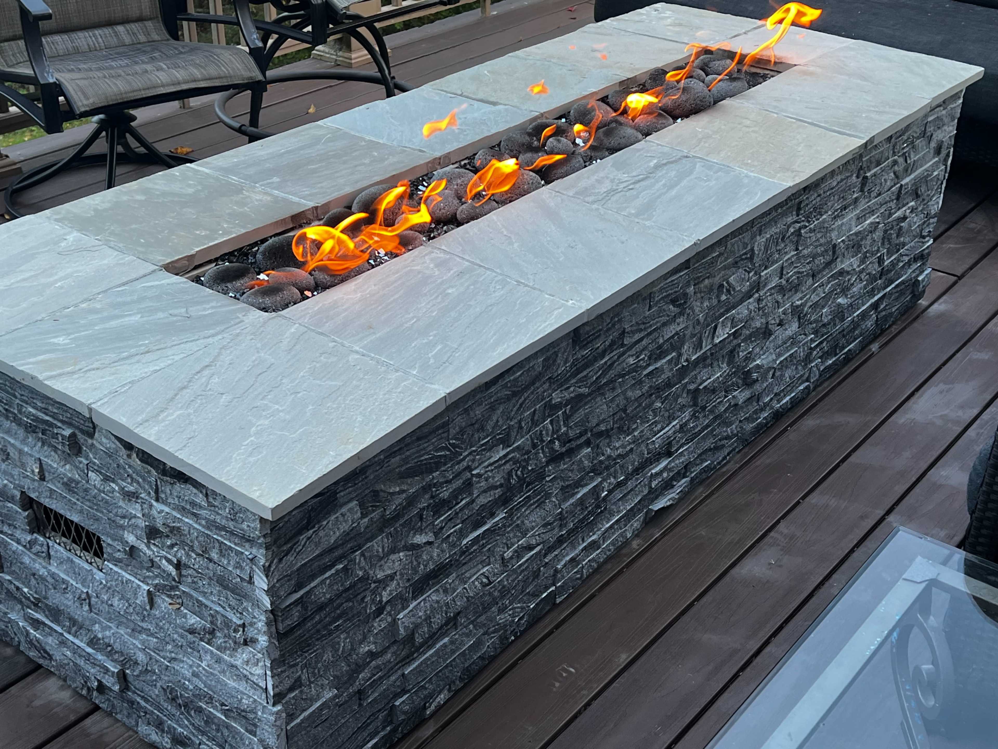 A rectangular stone fire pit with flames and decorative rocks is positioned on a wooden deck.
