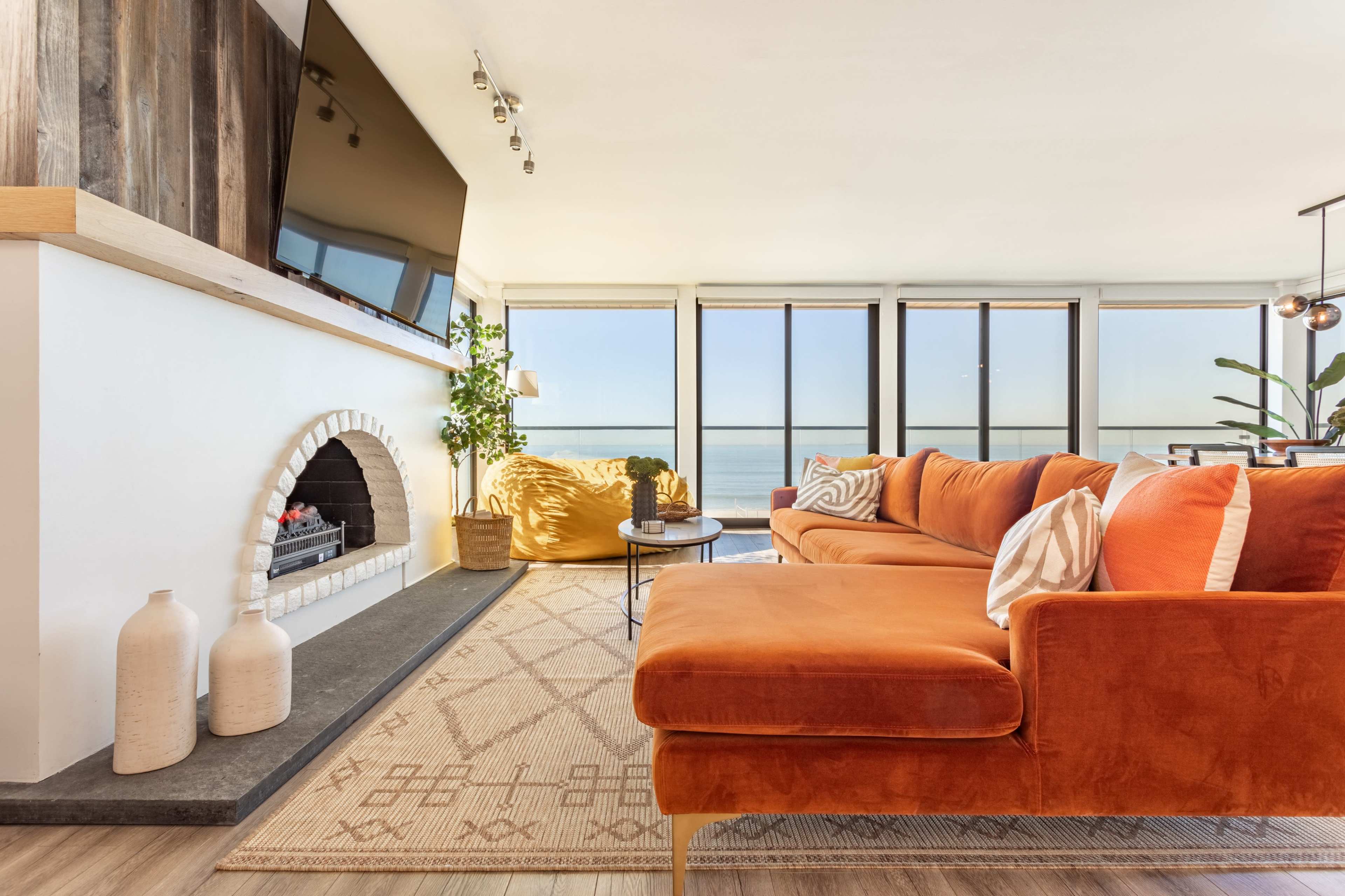 A modern living room features an orange sectional couch, a stone fireplace with a mounted television above it, and large windows overlooking a body of water.