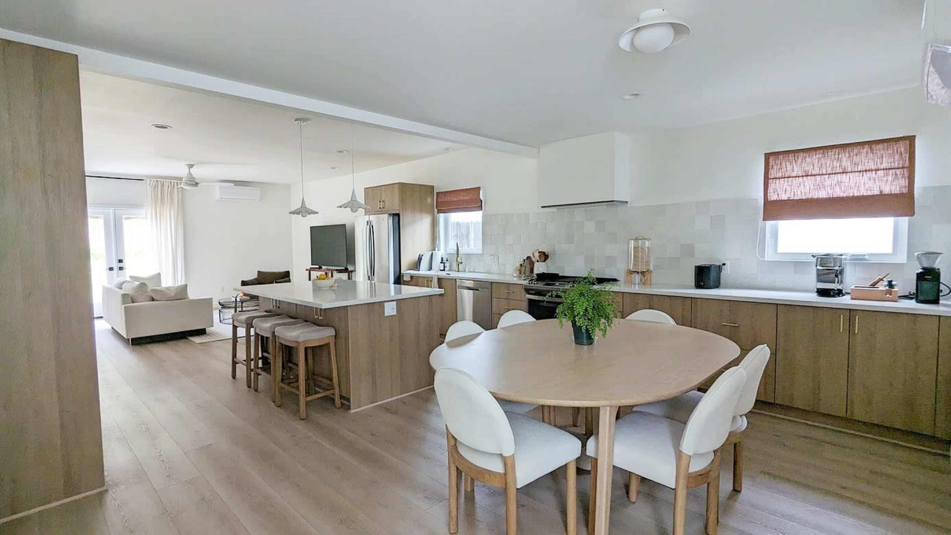 A modern kitchen and dining area feature light wood cabinetry, a circular dining table with six white chairs, and a living area visible in the background.