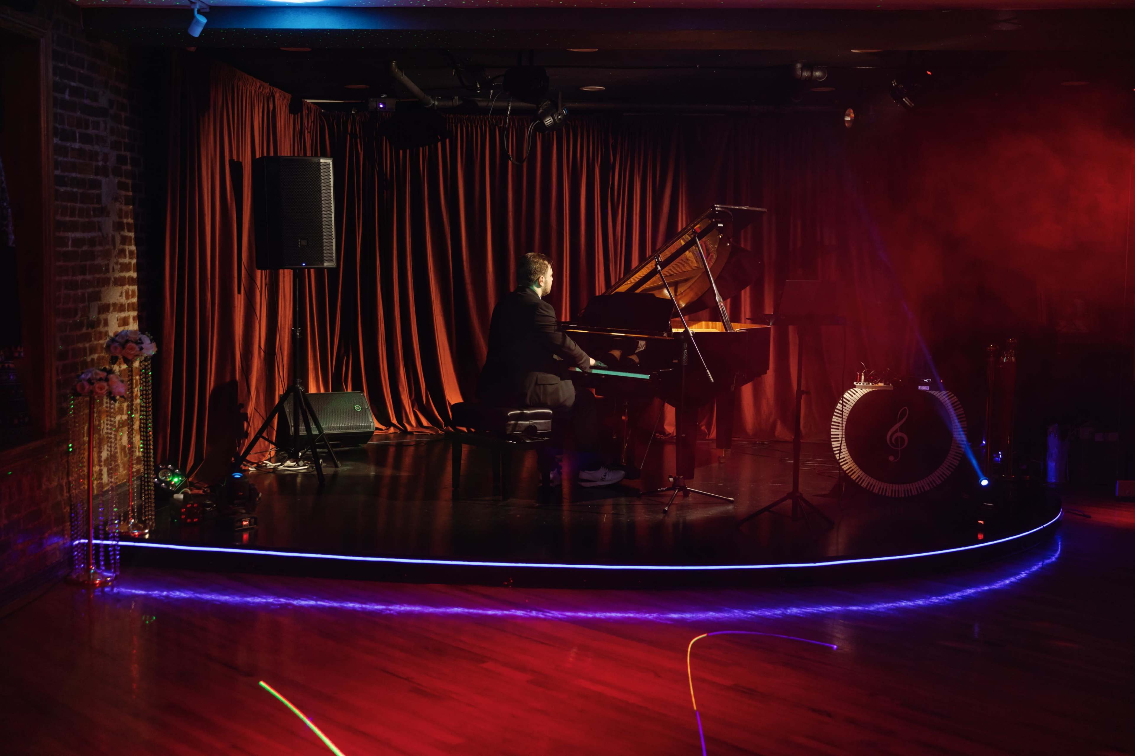 A pianist plays a grand piano on a stage with red curtains and colorful lighting in a dimly lit venue.