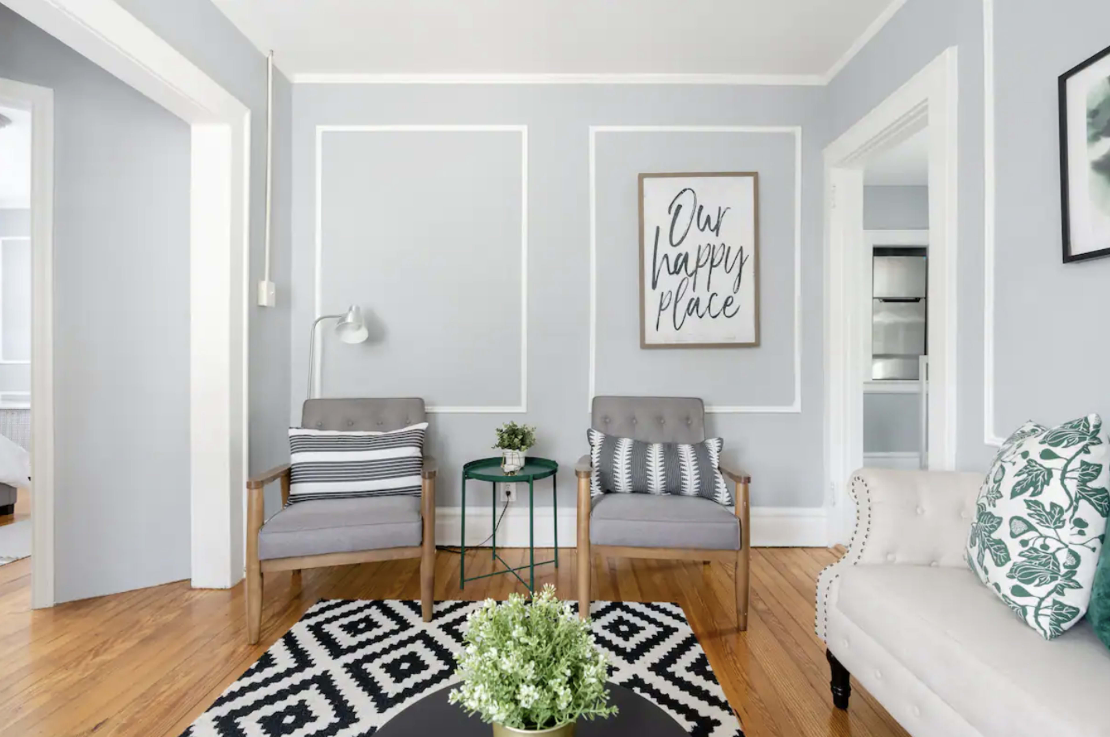 The image shows a living room with two gray armchairs, a small green side table, and a light-colored sofa, all set against light gray walls with decorative white framing.