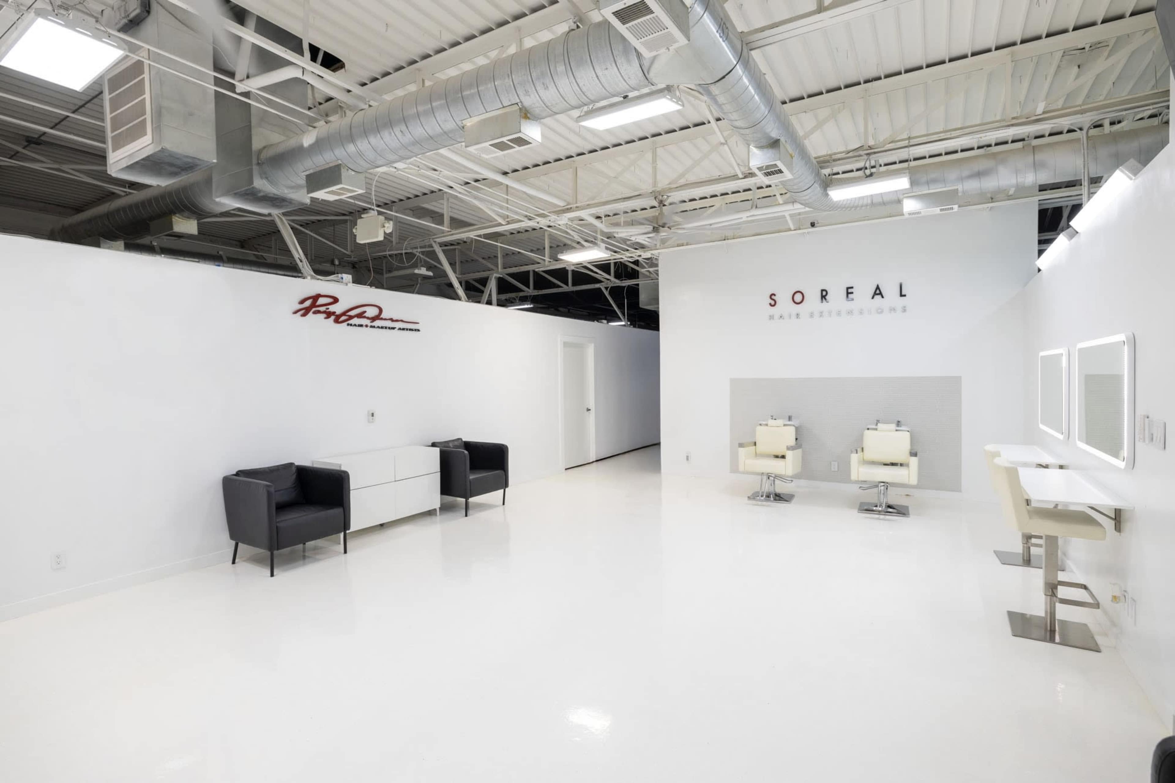 The image shows a modern, minimalistic salon interior featuring two styling chairs, a seating area with black chairs, and large mirrors on a white wall.