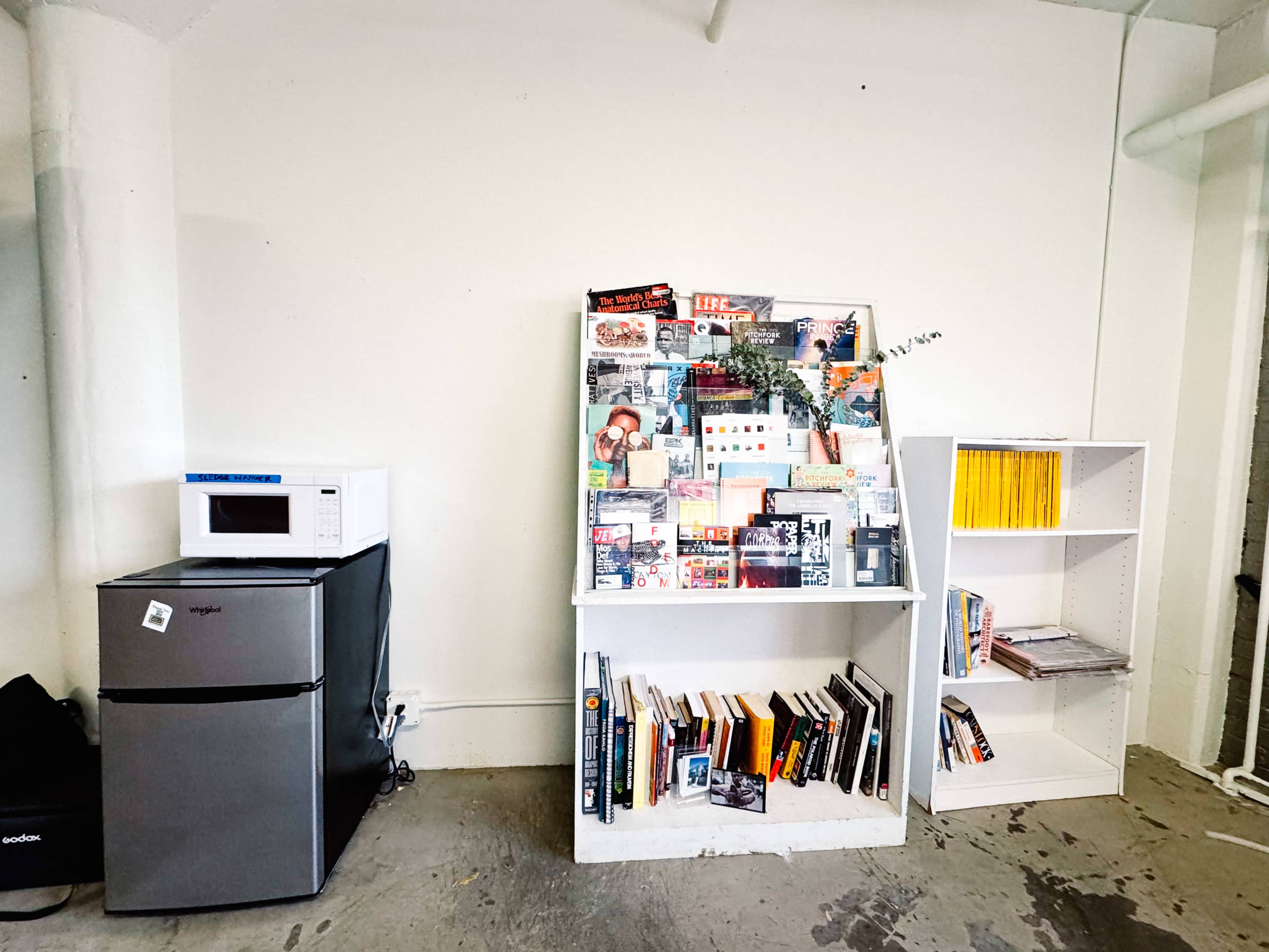 The image shows a room with a microwave and refrigerator on the left and two white bookcases displaying books and magazines on the right.