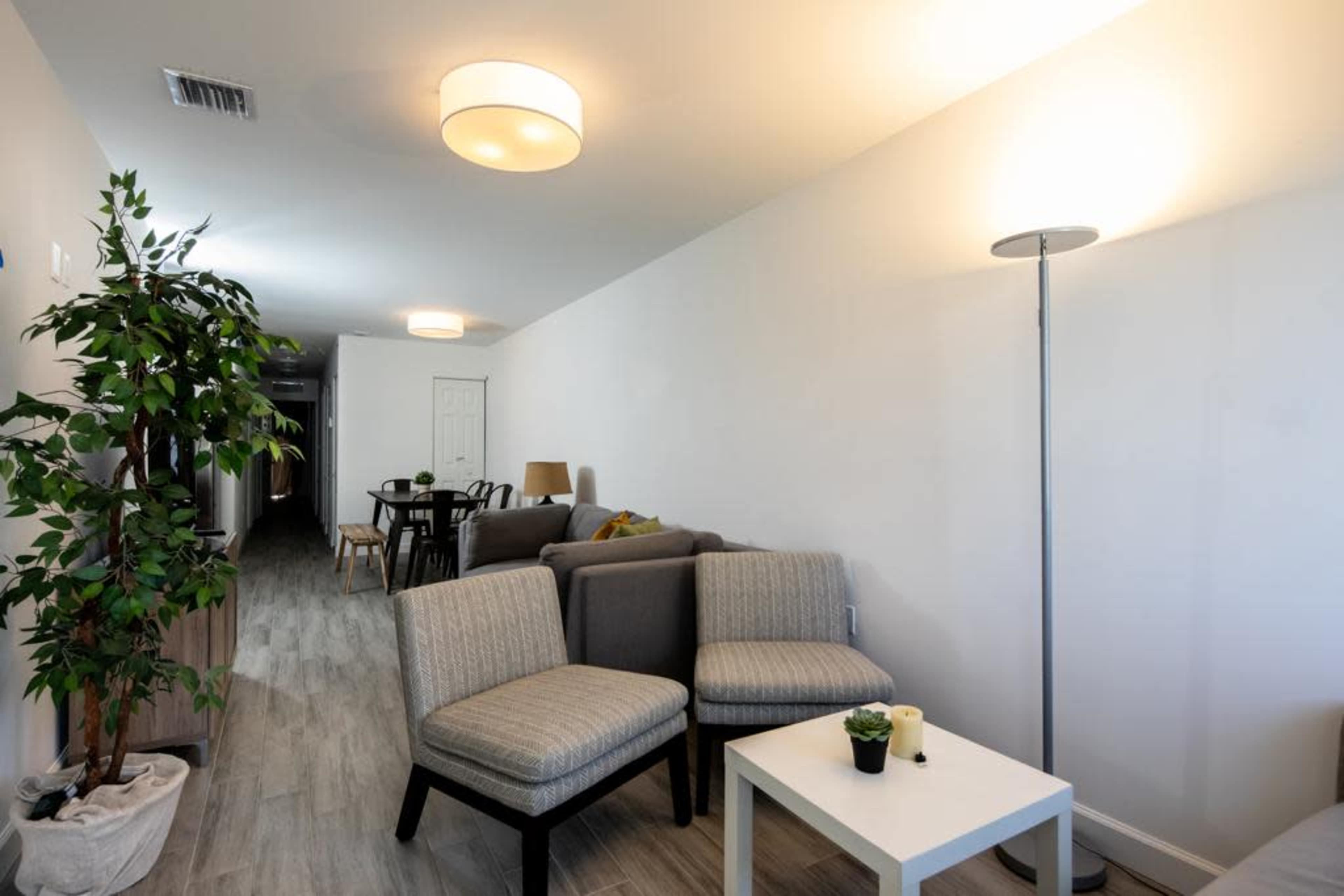 A modern, well-lit living space featuring a seating area with gray chairs, a small white table, and a floor lamp, along with a dining area in the background.
