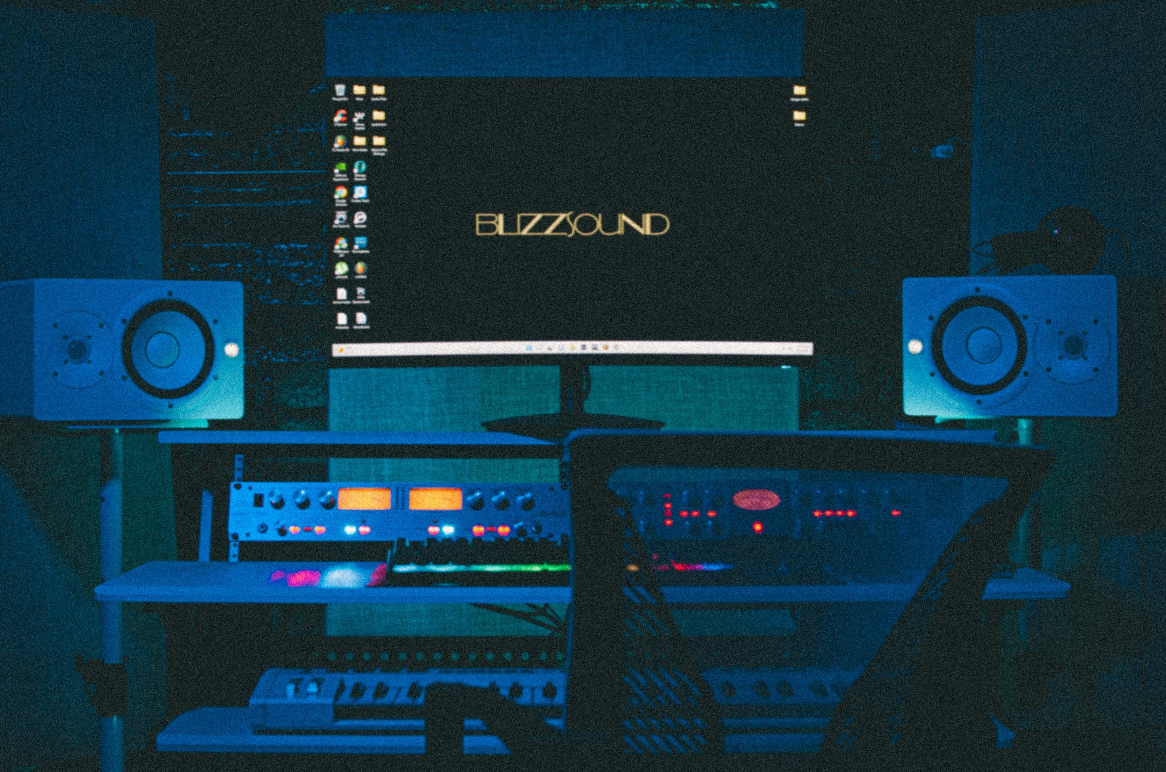 A home music studio features a large monitor displaying "BUZZSOUND," flanked by two audio speakers, with various music production equipment on the desk.