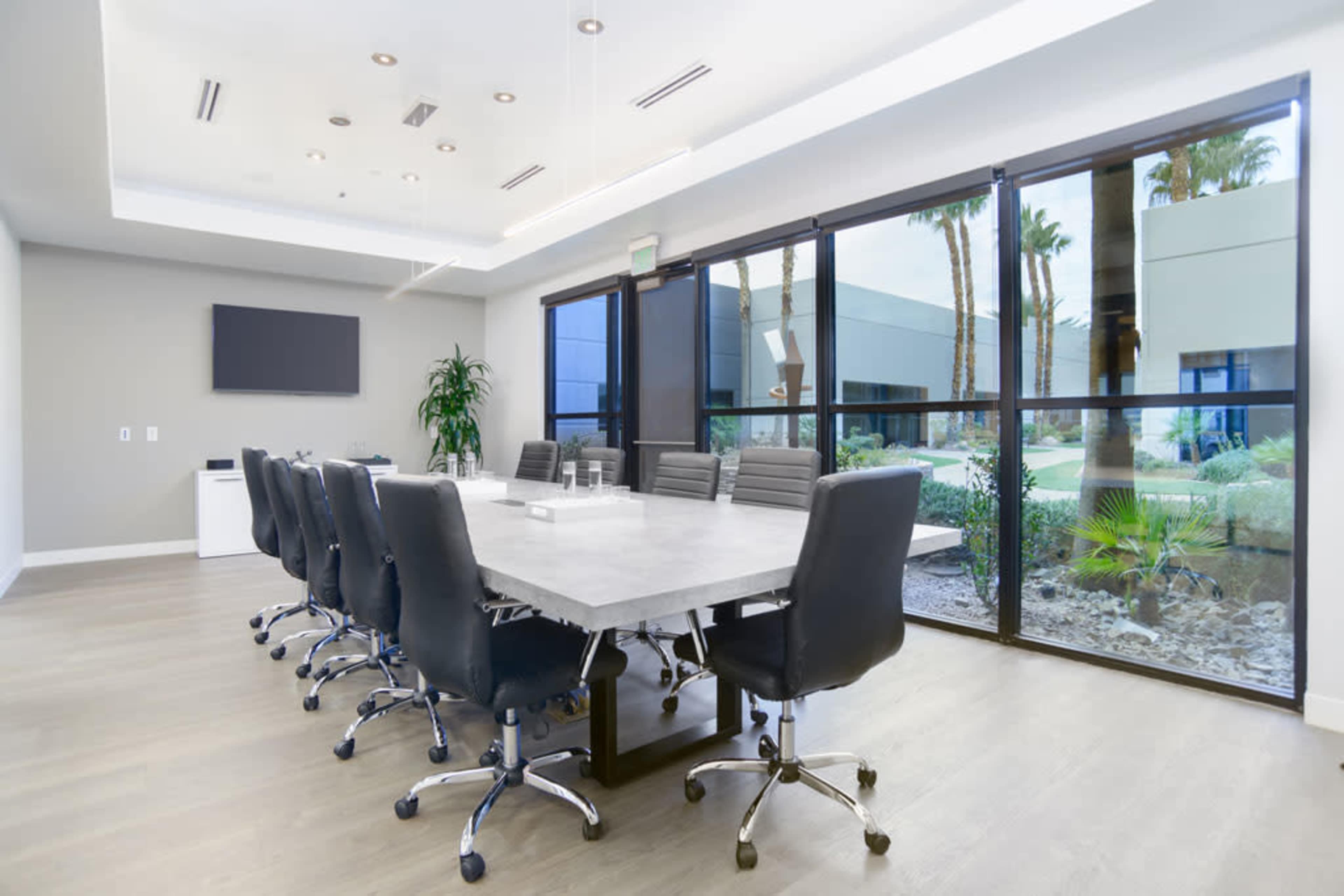 A modern conference room features a long table surrounded by black office chairs, with large windows overlooking a landscaped area.