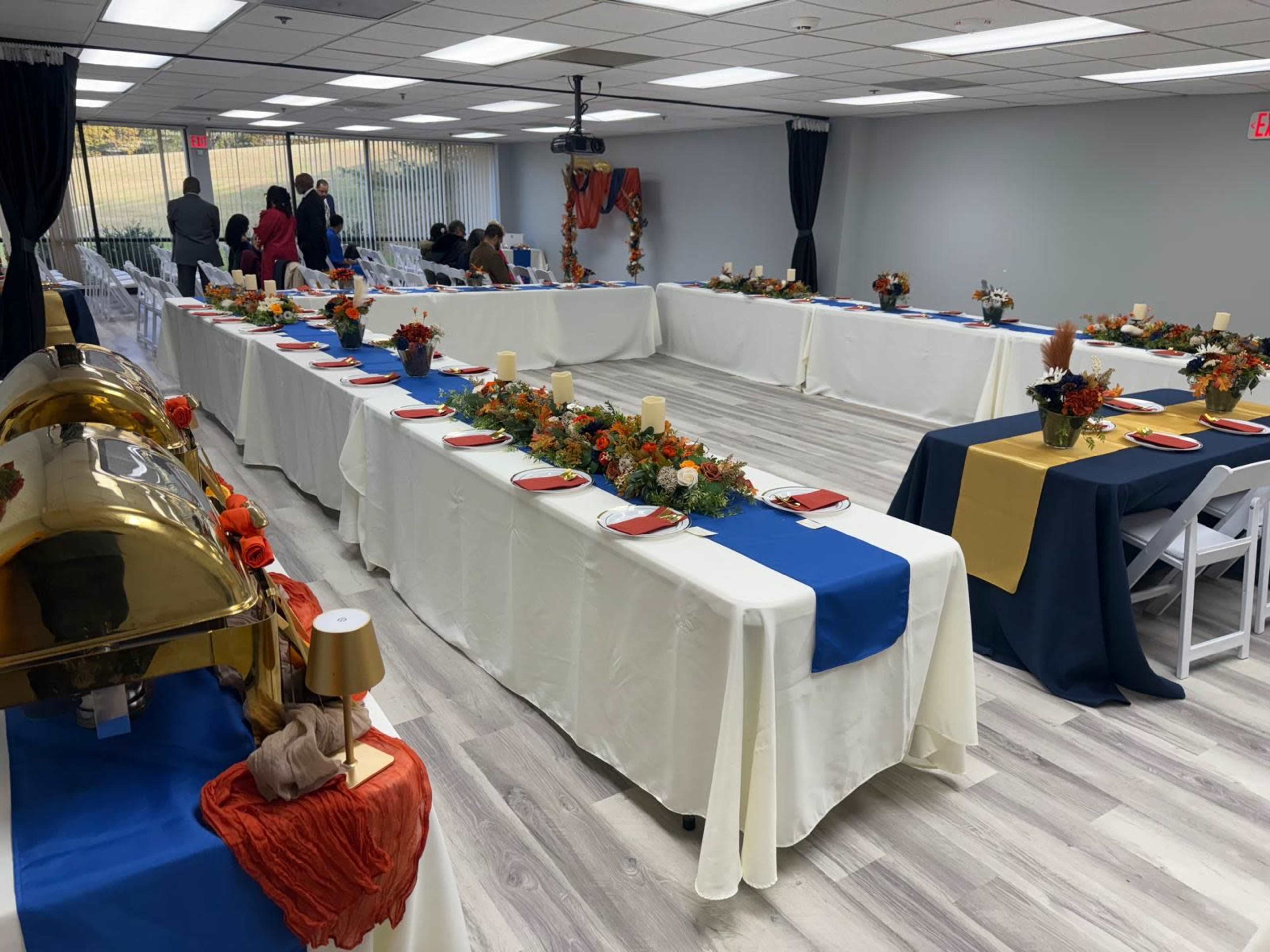 The image shows a spacious event room set up with long tables adorned with floral centerpieces and decorative tableware, all arranged for a gathering.