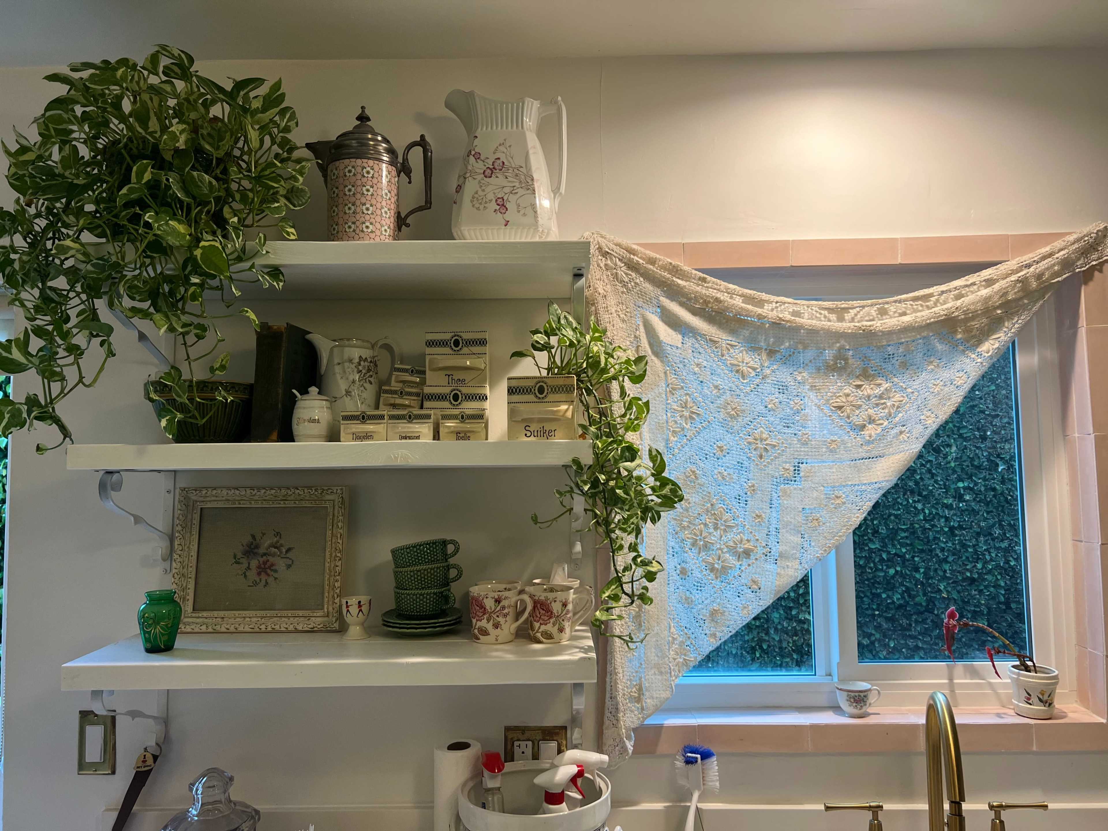 A kitchen shelf displays various dishes and a vintage teapot, accompanied by a green plant and a lace curtain covering a window.
