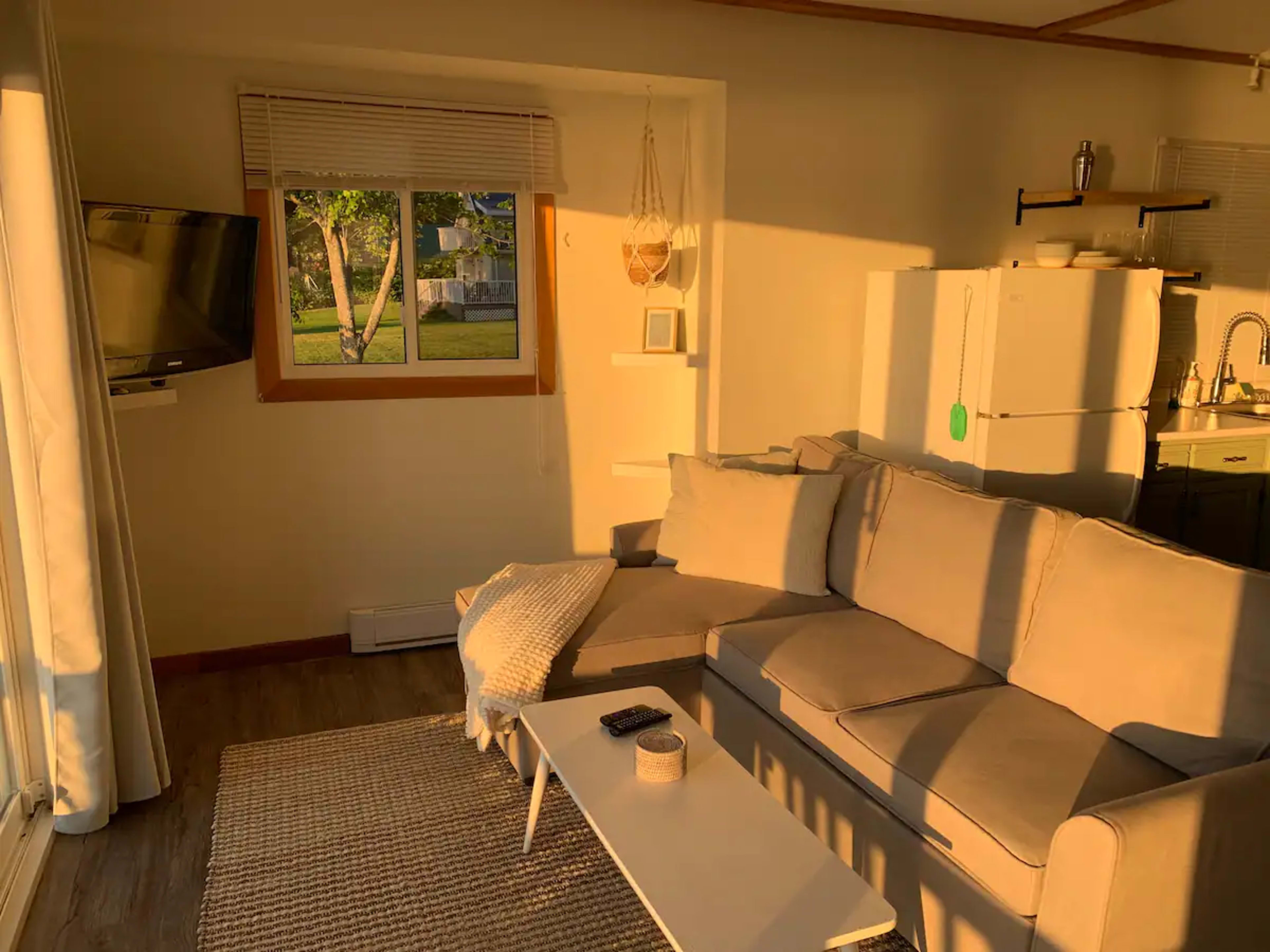 A well-lit living room features a beige sofa, a coffee table, and a window with sunlight streaming in.