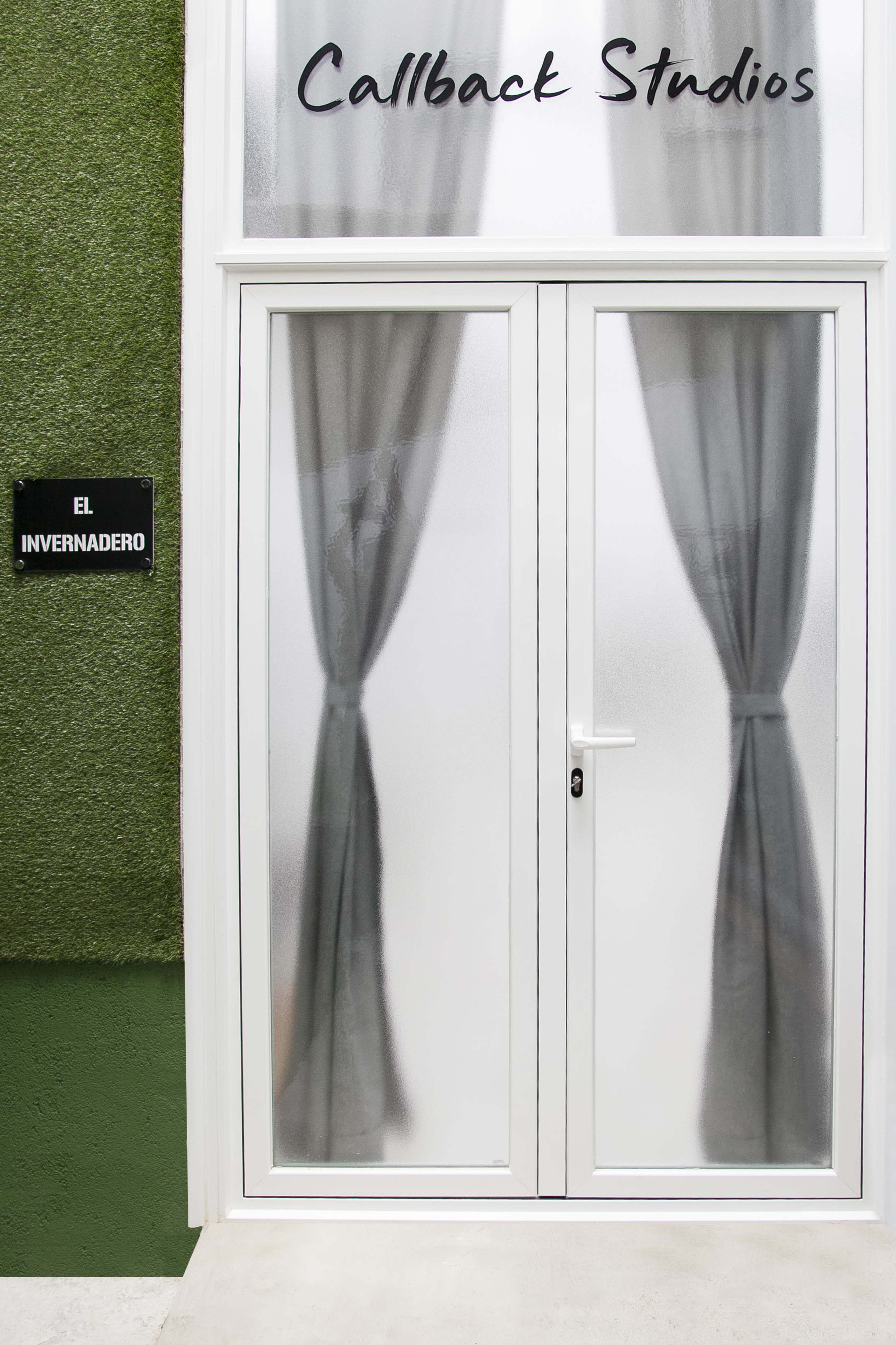 The image shows a frosted glass doorway with curtains and a sign reading "El Invernadero" next to the entrance of Callback Studios, set against a green wall.