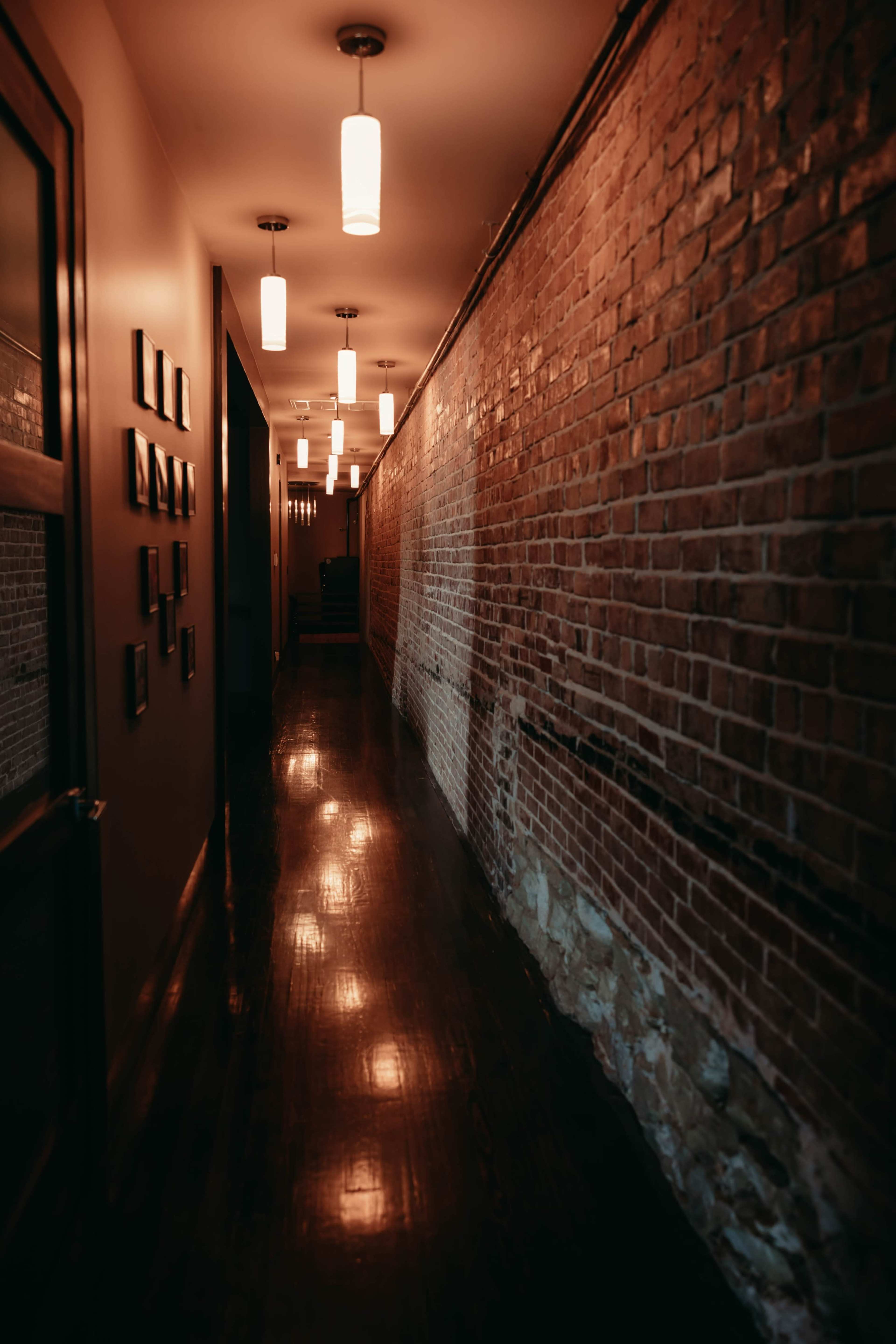 A narrow hallway with exposed brick walls and pendant lights reflects a polished wooden floor.
