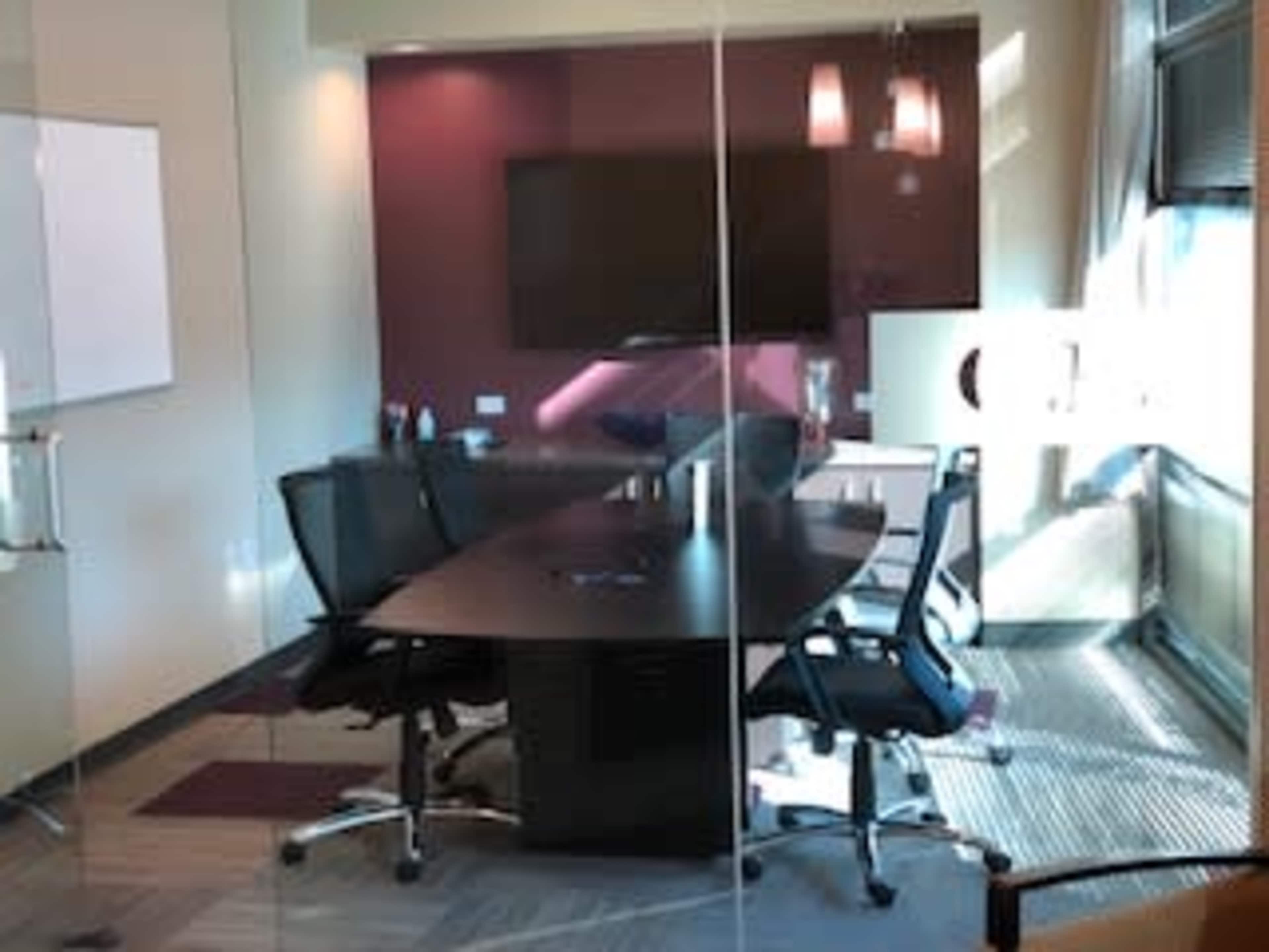A modern conference room with a round table, four black chairs, a large screen on the wall, and a glass wall.