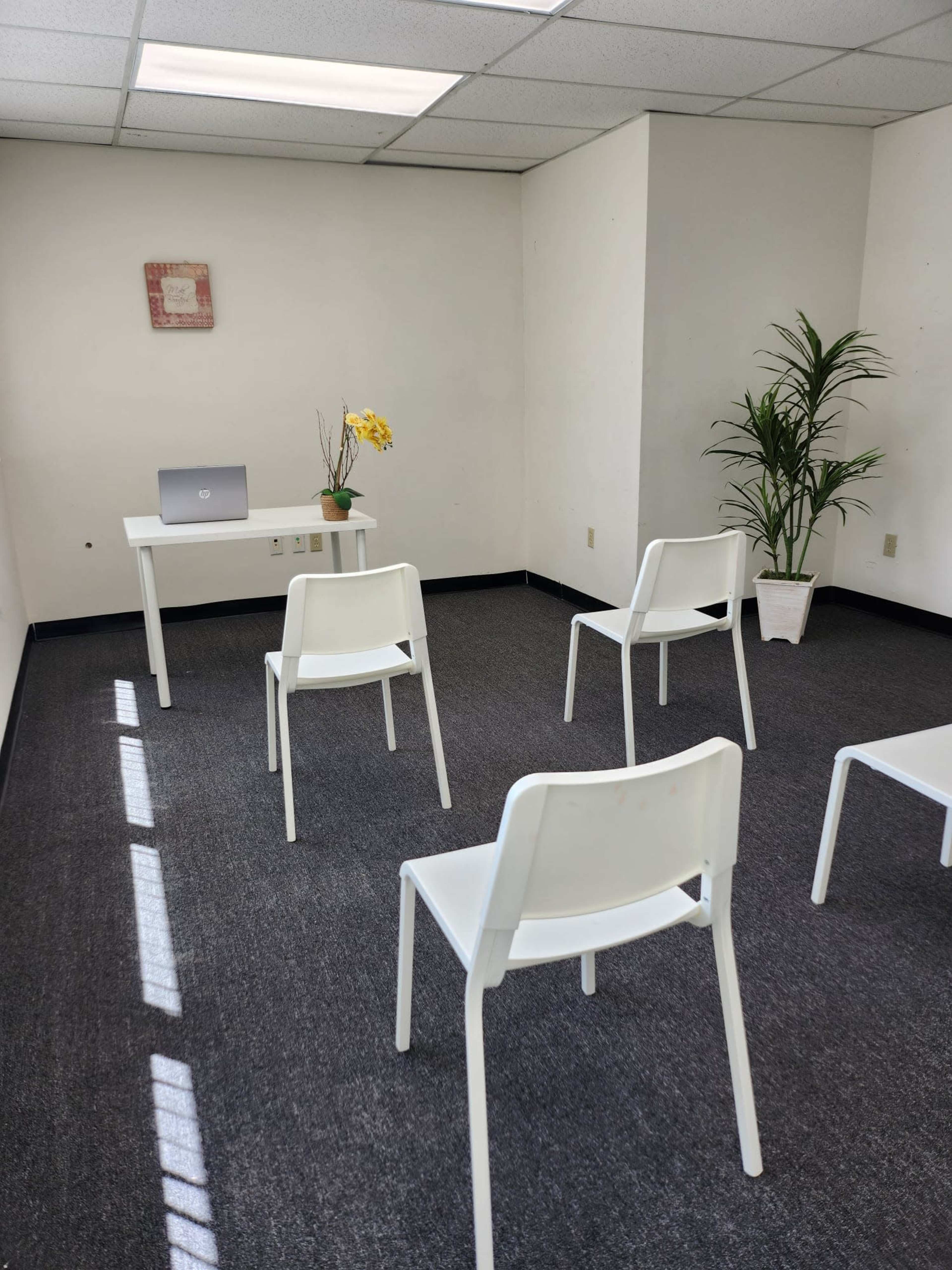 A sparsely furnished room features a table with a laptop, a plant in a pot, and four white chairs arranged around the space.