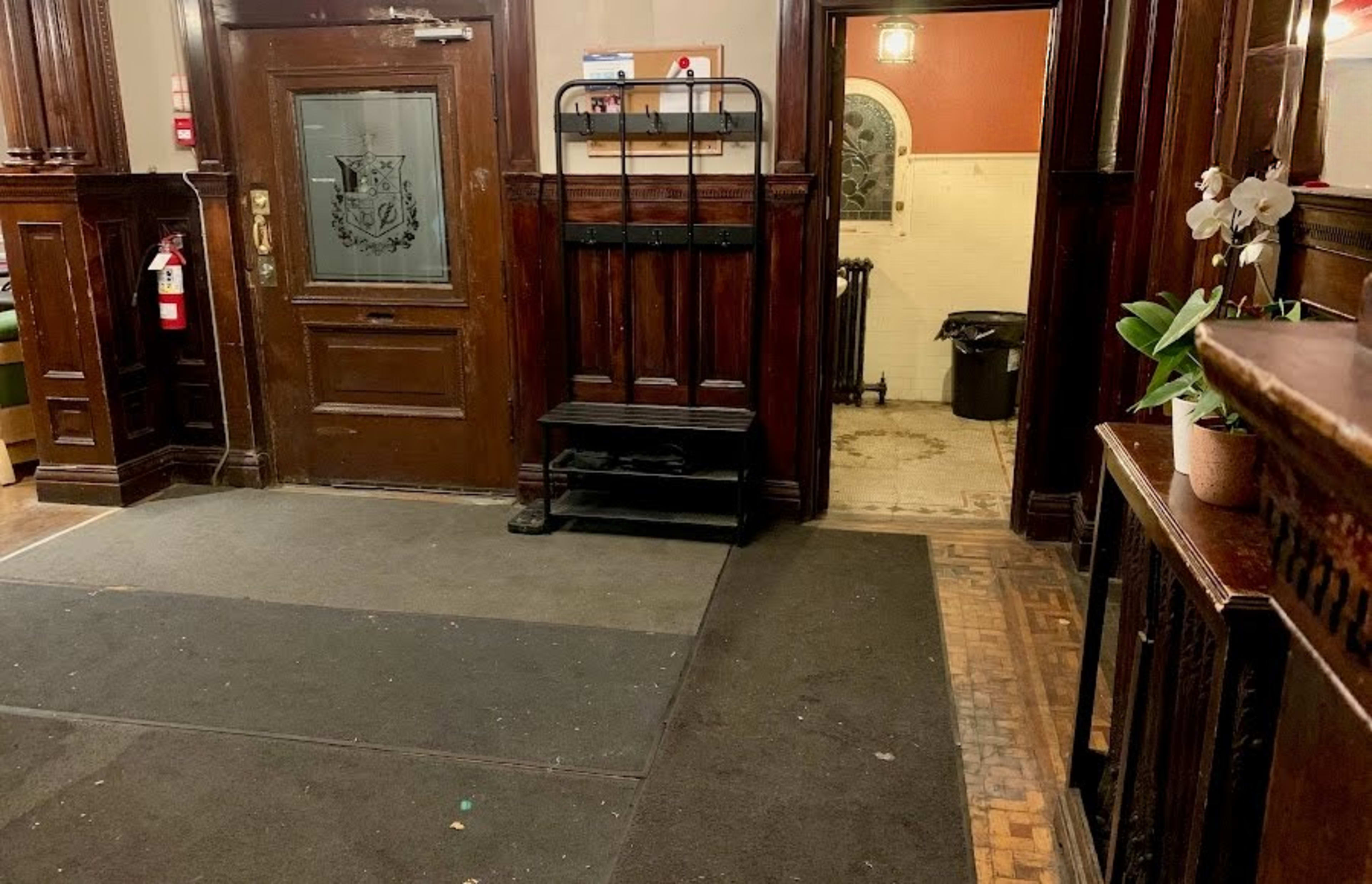 The image shows an interior entrance area with wooden paneling, a coat rack, and a visible doorway leading to a room with tiled flooring.
