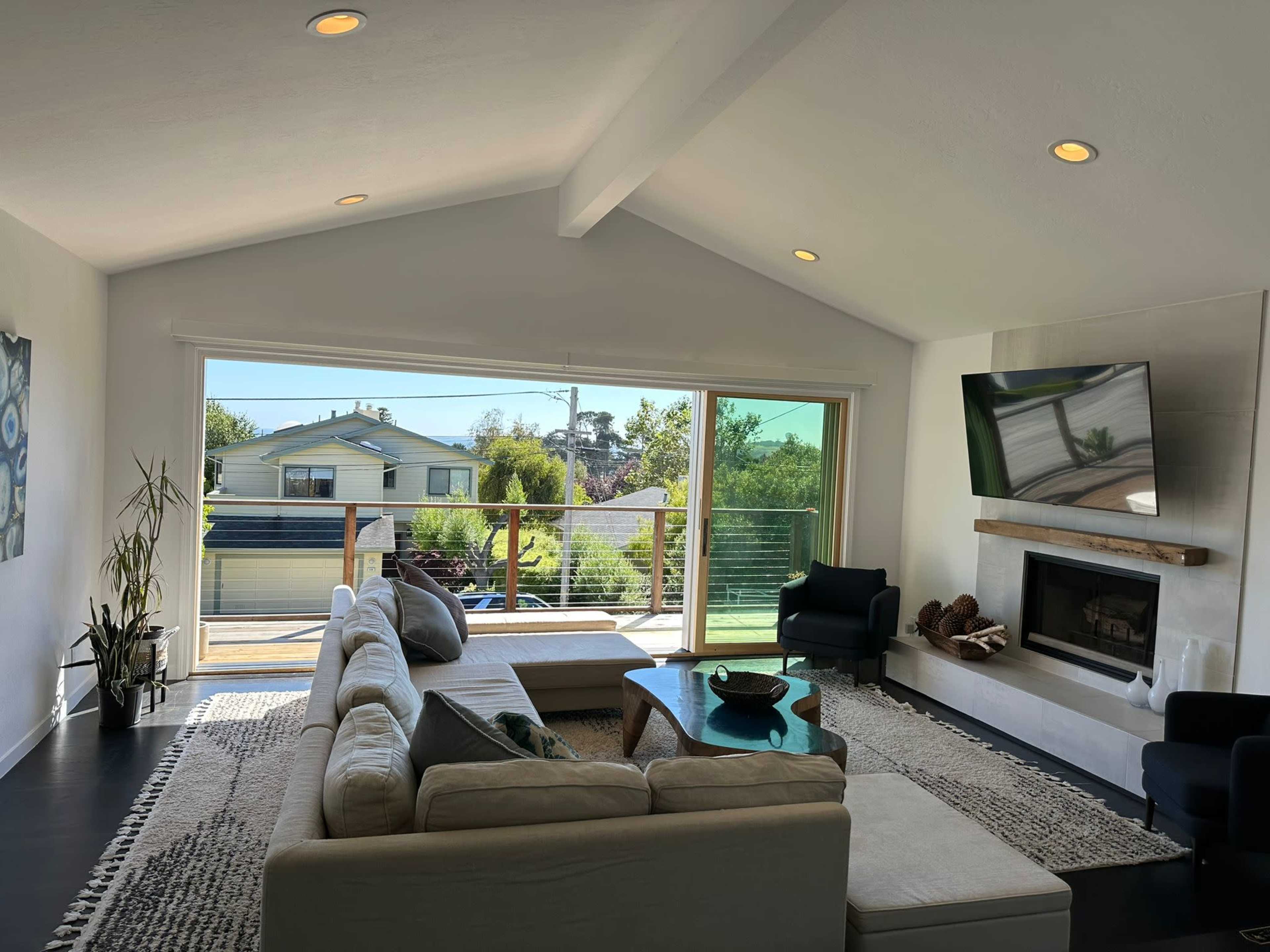 The image shows a modern living room with a sectional sofa, a coffee table, and a large window overlooking a patio and garden area.