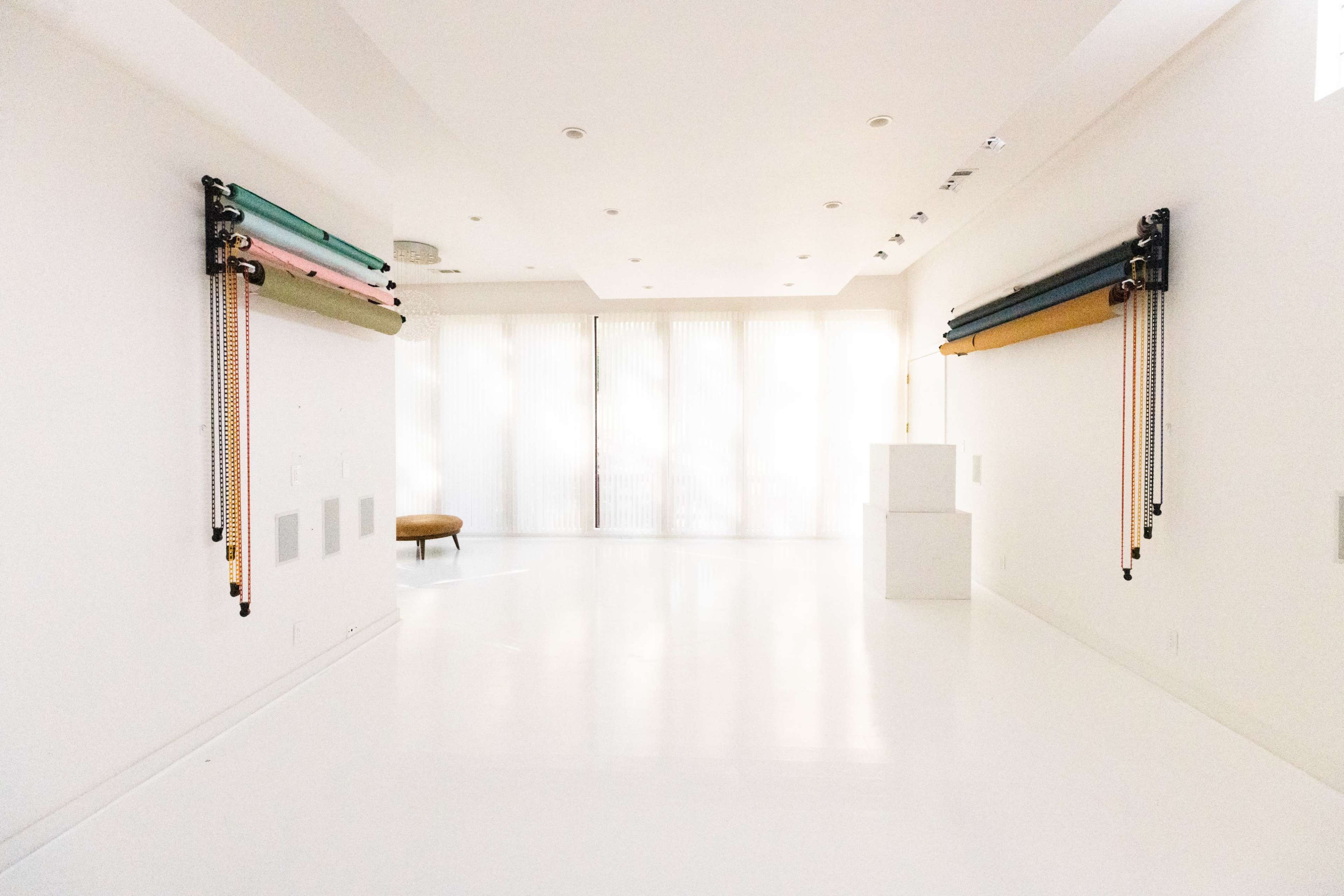 A minimalist room with white walls and flooring features two sets of colored fabric rolls mounted on the walls and large windows with sheer curtains.