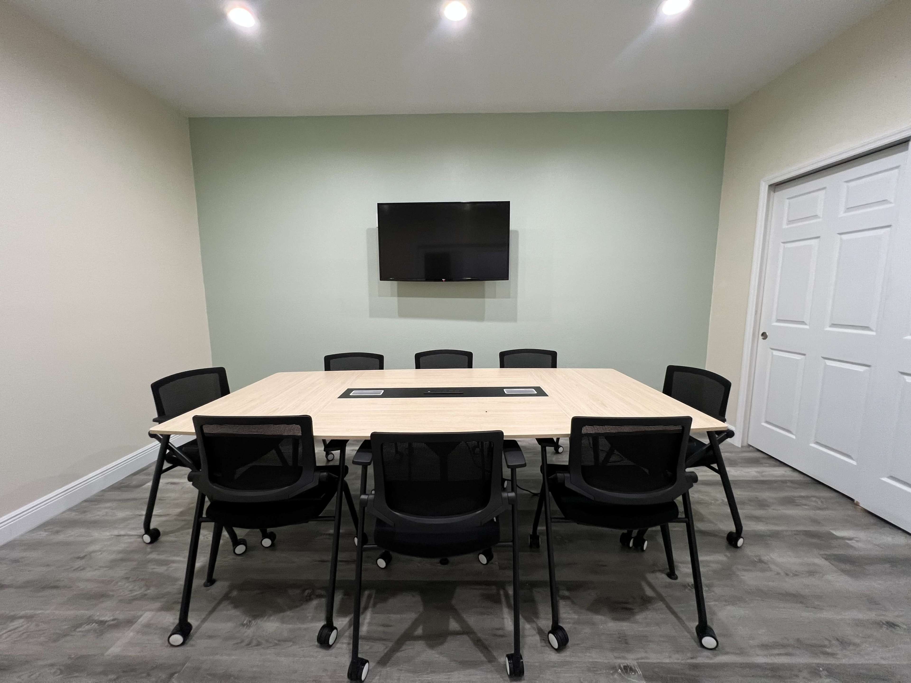 The image shows a conference room with a rectangular table surrounded by black chairs, a mounted television on the wall, and a closed door.
