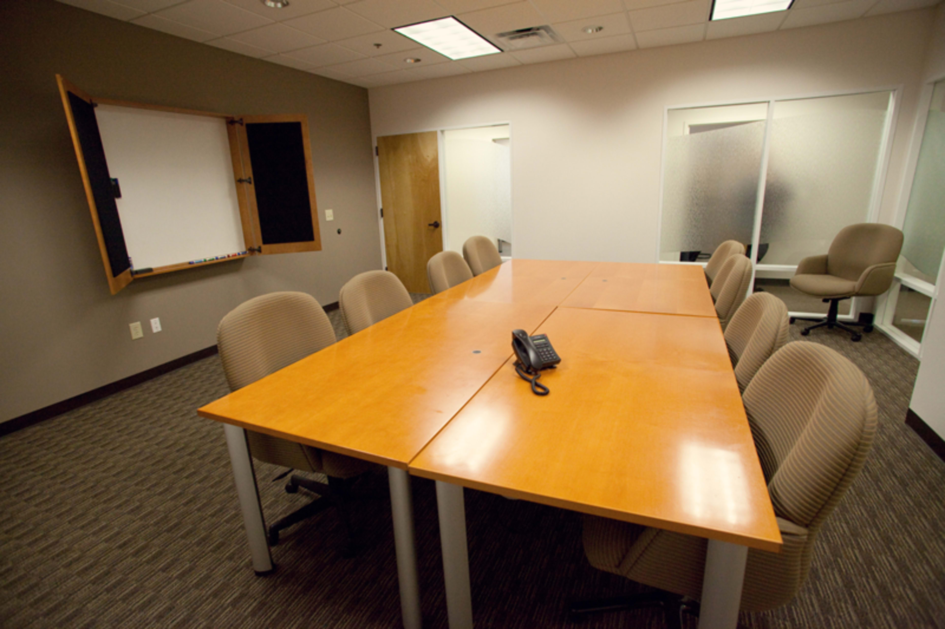 A professional conference room featuring a large wooden table surrounded by several beige chairs, with a whiteboard mounted on the wall and a phone placed in the center of the table.
