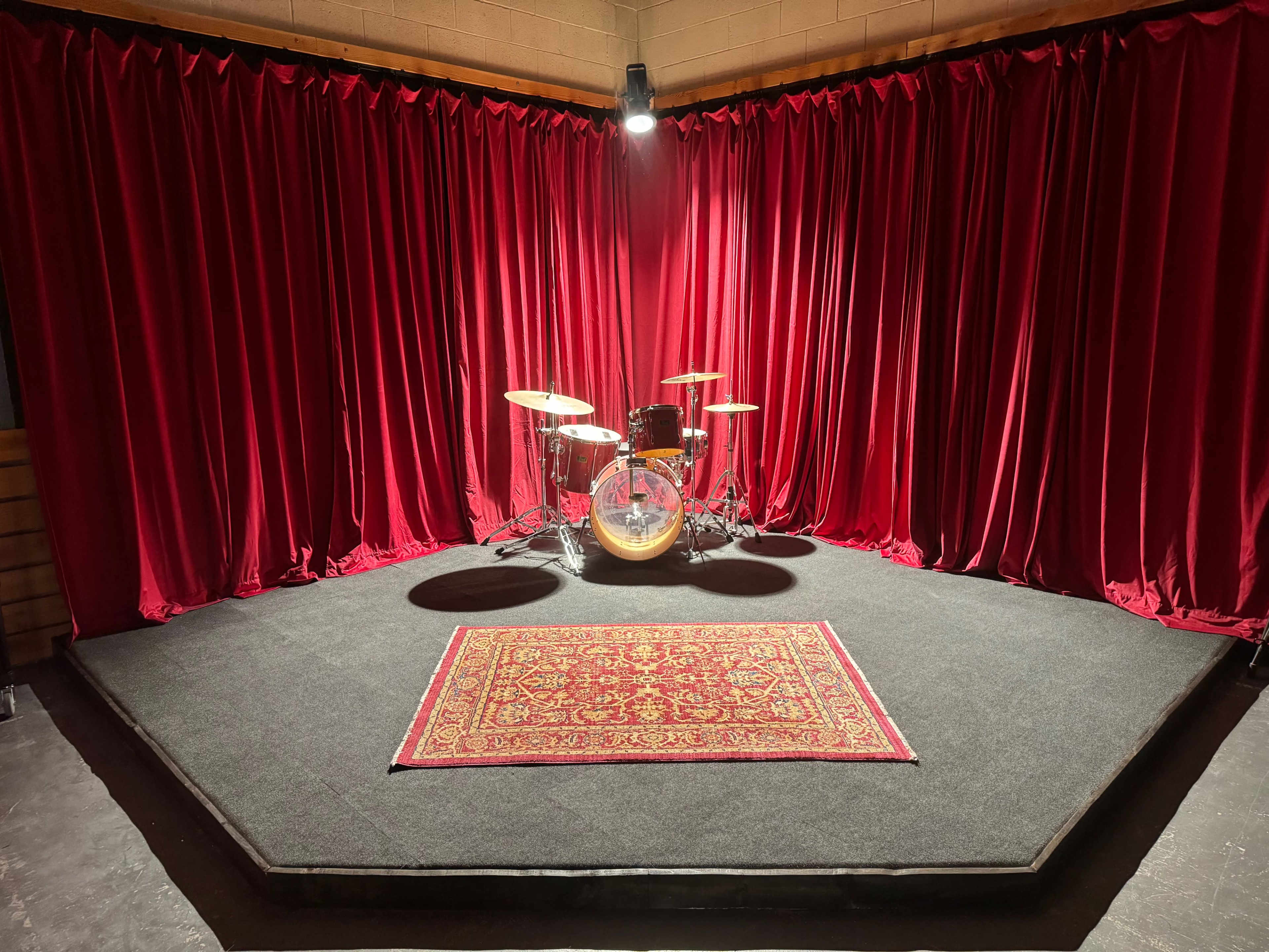The image shows a music stage with a drum set set against a backdrop of red curtains and a patterned rug on the floor.