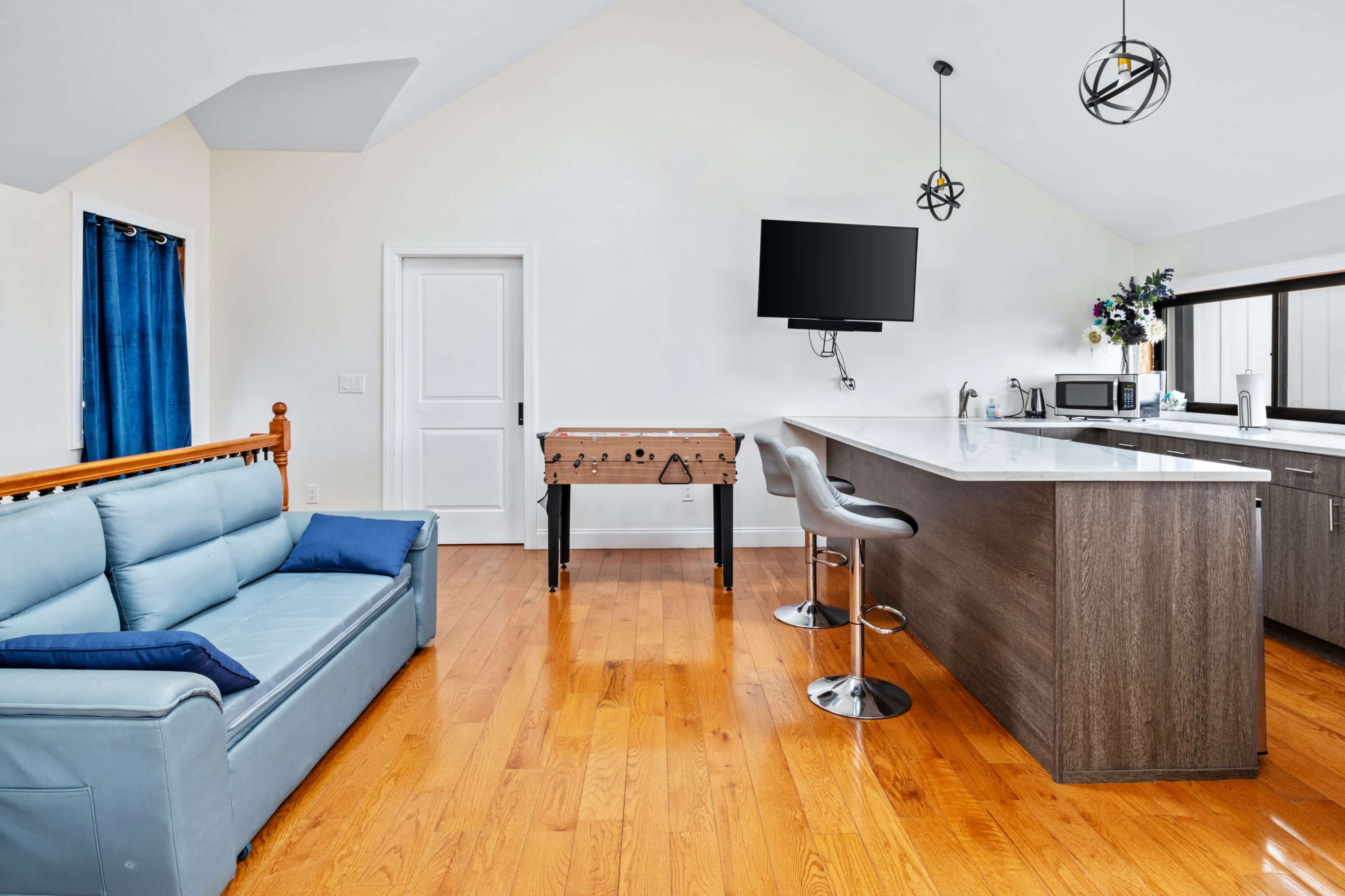 The room features a modern kitchen area with a bar counter, a foosball table, and a couch, all illuminated by natural light from large windows.