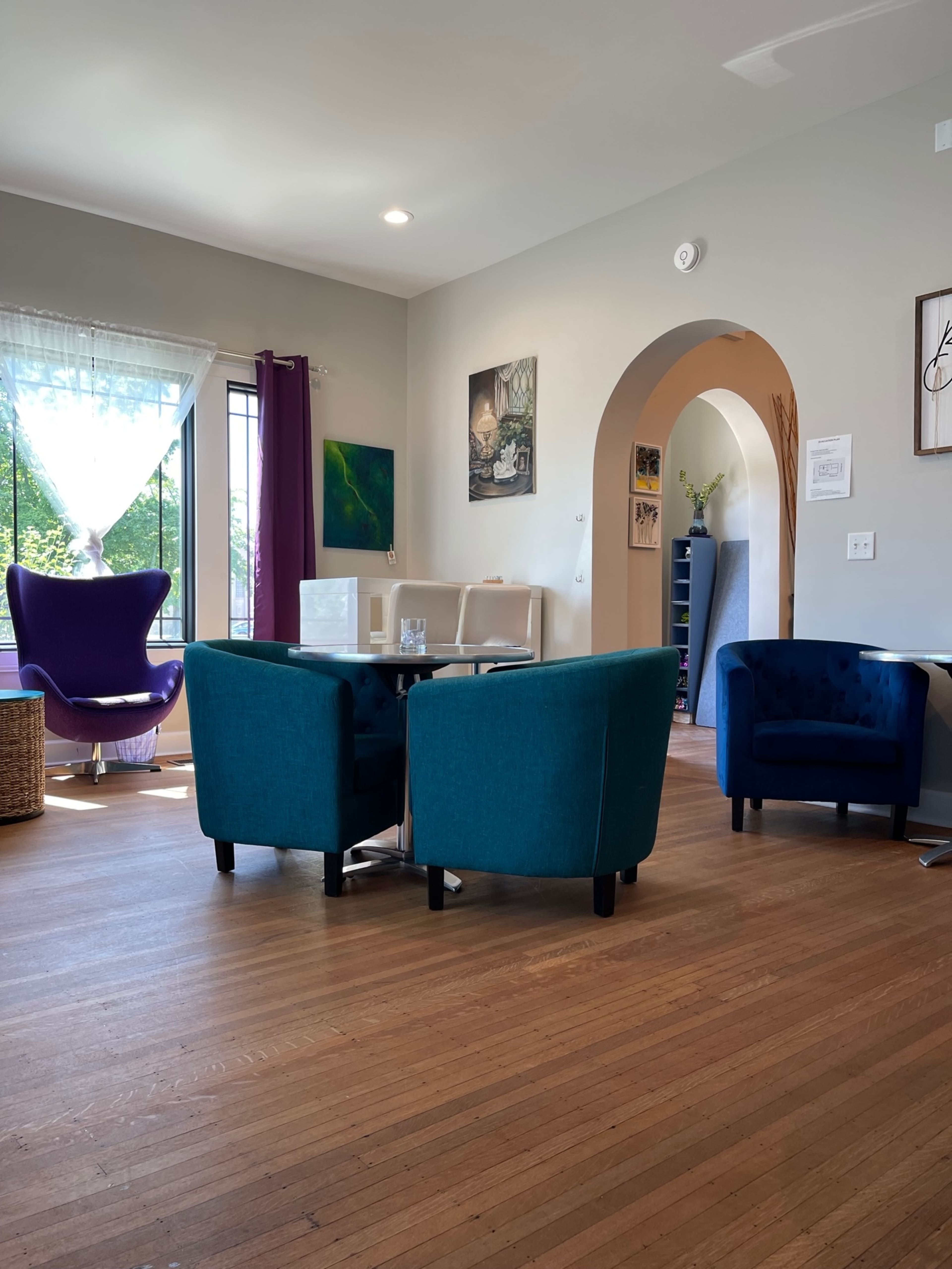 A cozy indoor space featuring two teal armchairs and a glass table, with a purple chair in the background and large windows allowing natural light to enter.